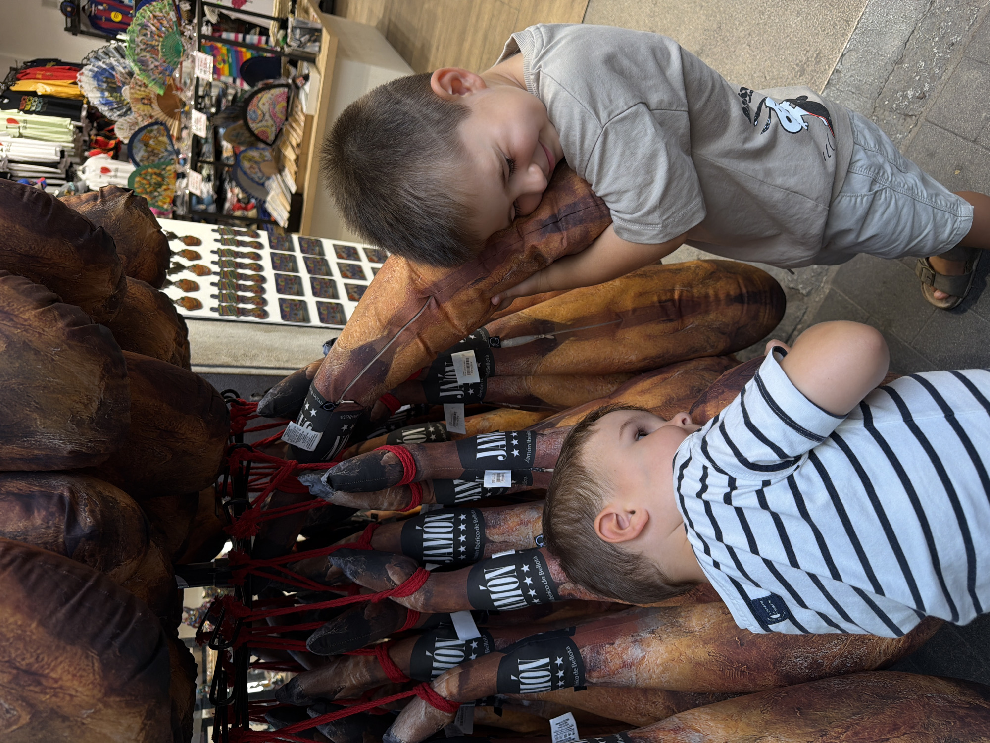 Two young boys are in a market stall, surrounded by large hams hanging from a rack. The boy on the right is wearing a light gray t-shirt and shorts, with sandals. He is hugging one of the hams and has his eyes closed, with a smile on his face. The boy on the left is wearing a striped t-shirt and is looking towards the hams. The background shows shelves stocked with various souvenirs, including fans and t-shirts. The lighting suggests it is daytime.