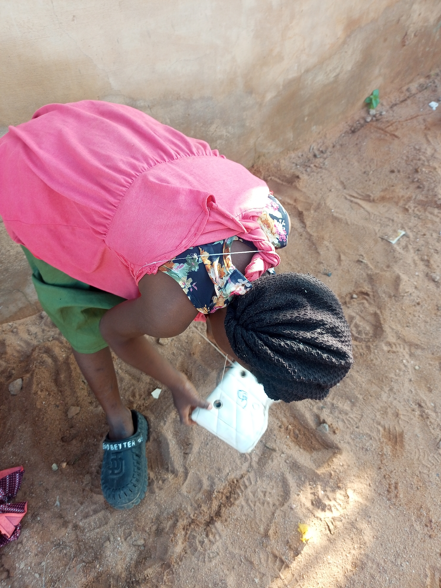 A person is bending over in a sandy area. The person is wearing a pink shirt, a floral-patterned top, a green skirt, and a black shoe on their left foot. The person is holding a white bag with a blue design in their right hand. The background consists of a textured wall and sandy ground with some small plants and debris. The scene appears to be outdoors in a dry, possibly rural or undeveloped area. The location in Tempure, Nigeria, is not discernible from the image itself.