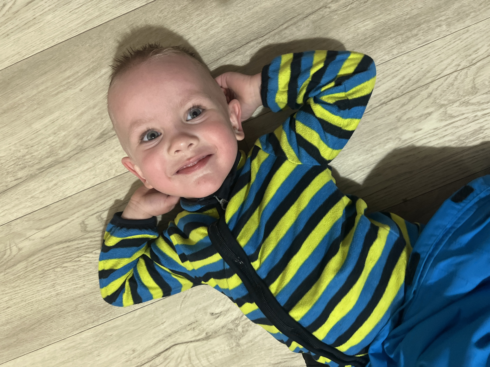 A young child, possibly a toddler, is lying on a wooden floor. The child is wearing a striped top of yellow and blue and blue trousers. The child is smiling and has their hands behind their head. The location is Марковский, Russia.