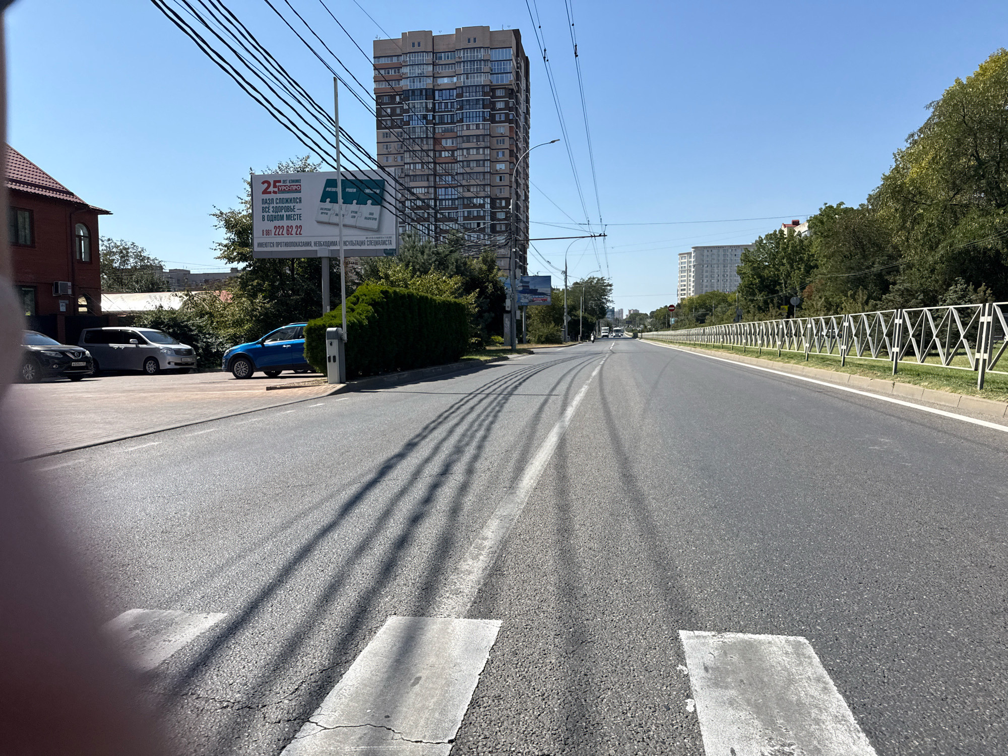 An eye-level, medium shot captures a street scene under a clear blue sky. In the foreground, a paved road with white pedestrian crossing markings dominates the lower portion of the frame. To the left, a red-brick building with an arched window is visible, and parked cars are present. A billboard stands near the building. Further down the road, several tall apartment buildings and trees line the street. The perspective is from the street, looking forward.