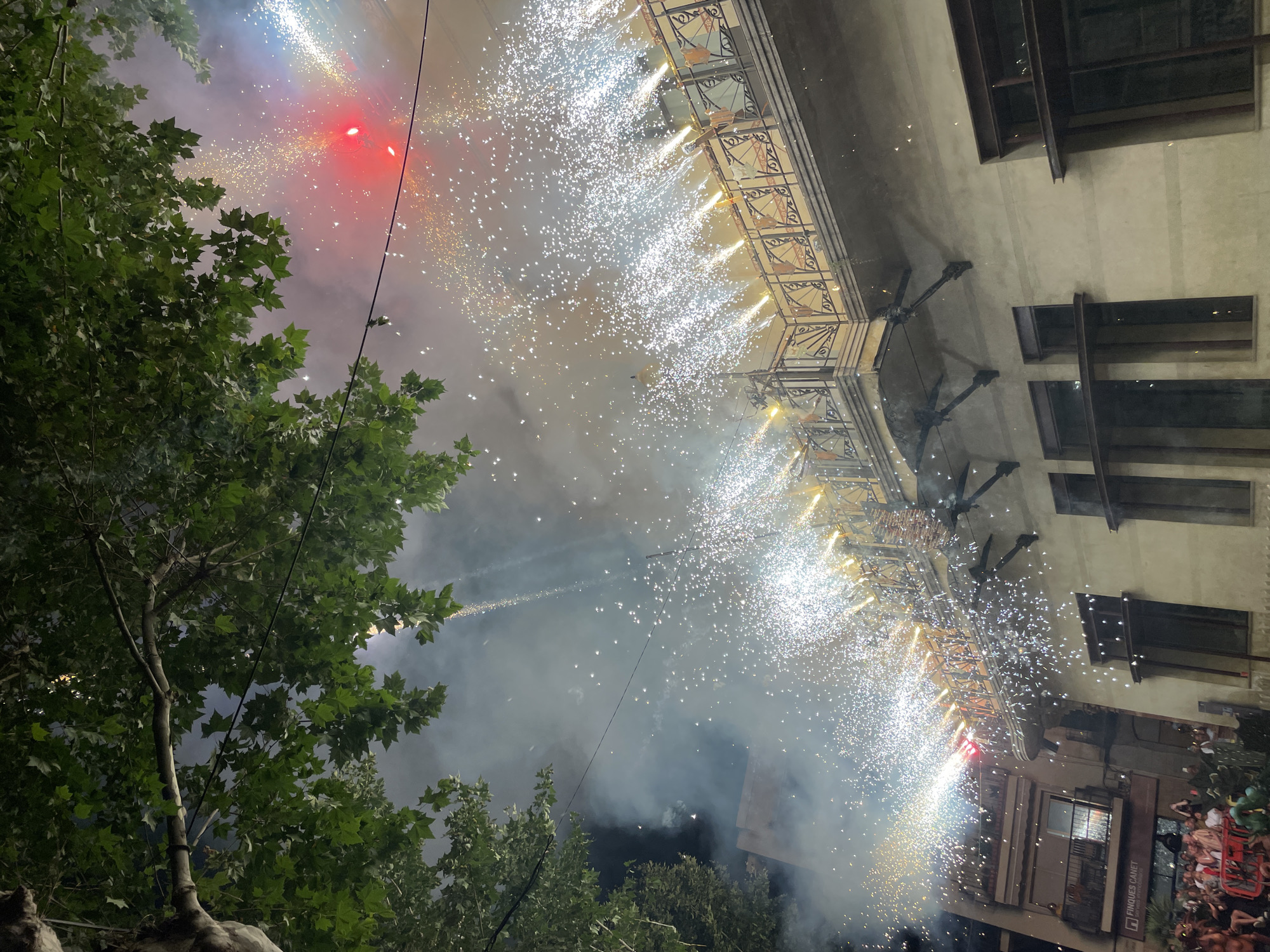 The image depicts a nighttime scene in Canet de Mar, Spain, characterized by a fireworks display. In the foreground, lush green tree branches frame the upper portion of the image. Below, a building with a decorative balcony is illuminated by the pyrotechnics. Fireworks are erupting from the balcony railing, creating a shower of sparks and smoke that cascades downwards. Some fireworks are also visible higher in the sky, including a bright red one. The building itself is light-colored with dark-framed windows. To the left, a crowd of people can be seen gathered on a street, some appearing to be watching the spectacle. A sign reading "FINQUES CANET" is visible on a building to the far left. The overall atmosphere suggests a festive event or celebration.