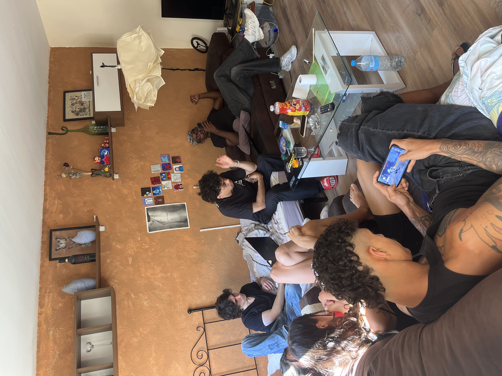 A rotated, high-angle shot depicts a room with an orange textured wall on the left, and wooden flooring. Several people are congregated within the space, some lounging on a brown couch and a bed covered with a light-colored blanket. One individual is focused on a smartphone, displaying what appears to be a video game. Another person nearby also seems to be looking at the phone. A glass-topped coffee table cluttered with items like a water bottle, food container, and remote controls is visible. On the wall, various decorations are present, including framed pictures, small decorative tiles, and a wine rack. A television is partially visible in the background. There is no indication or sign that this image was taken in Barcelona, Spain.
