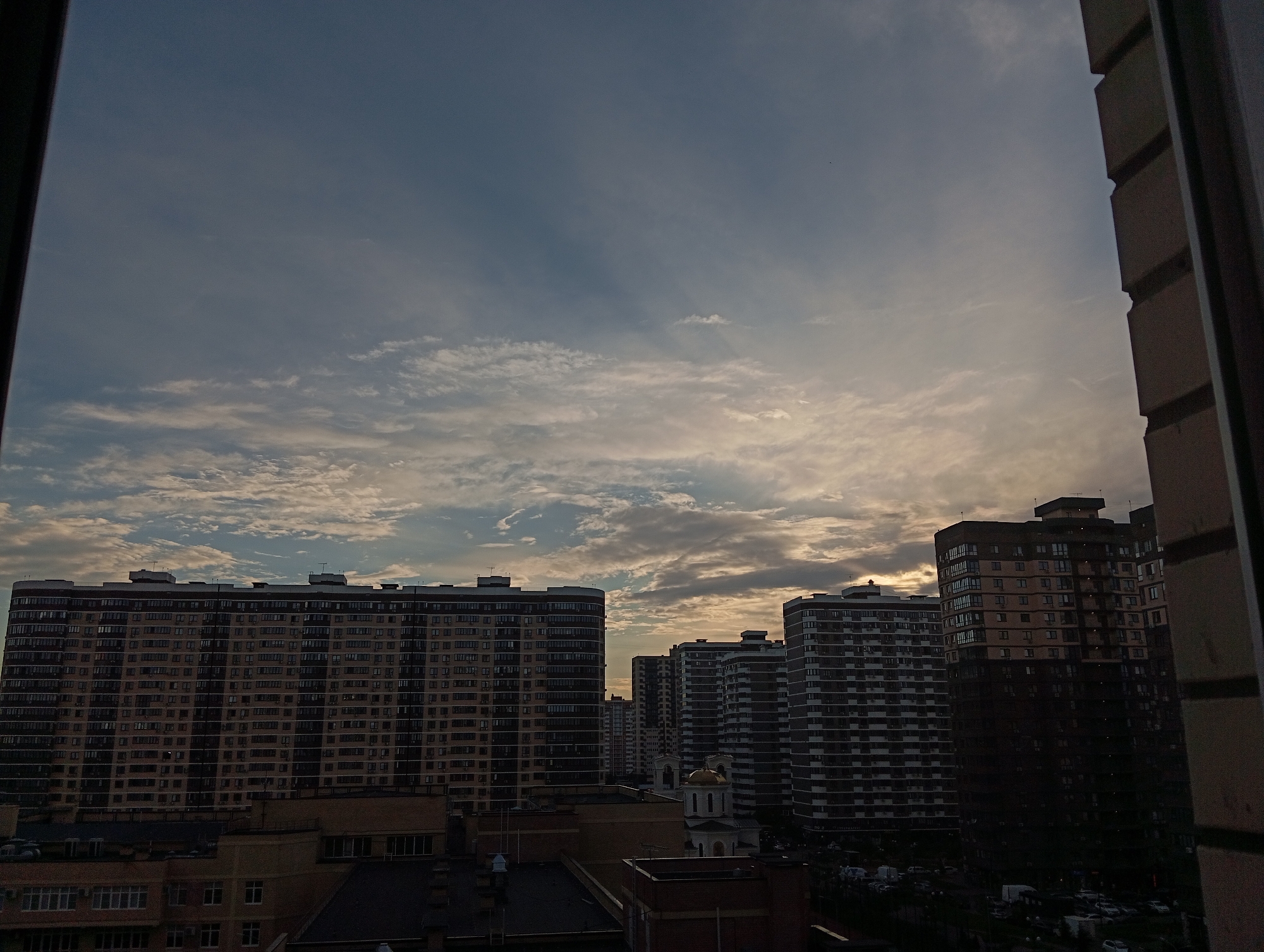 An eye-level, wide shot shows a cityscape with multiple high-rise apartment buildings under a cloudy sky. The sky is mostly blue with scattered white clouds, and there are some sun rays breaking through. The buildings are of varying heights and architectural styles, with many windows visible. In the foreground, a dark, brick-like structure frames the right side of the image, possibly a window frame or part of another building. The overall scene suggests an urban environment, possibly at dusk or dawn given the lighting.
