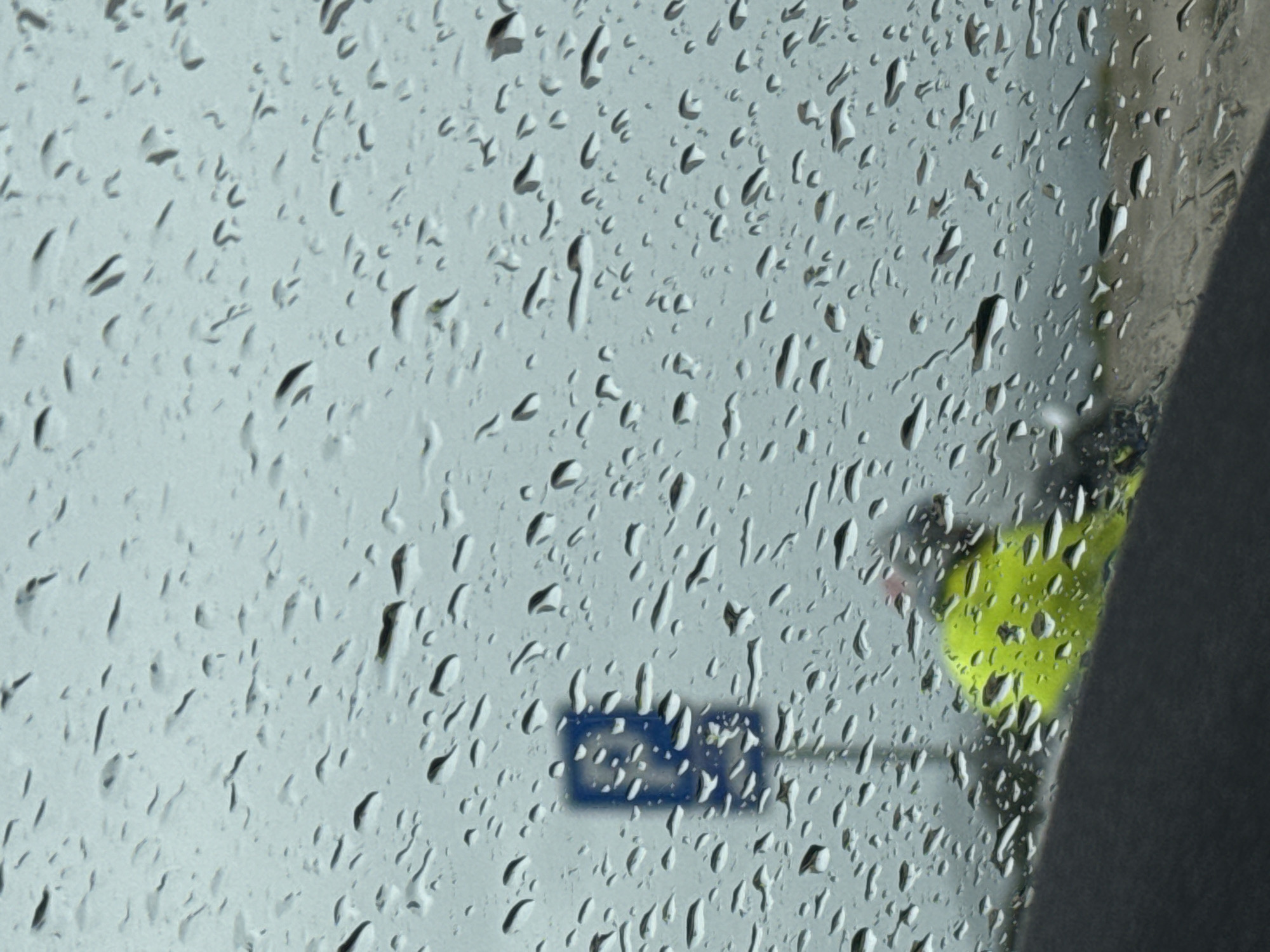 A blurred, wet window obscures the view of a street scene. Raindrops cover the glass. A blue sign is visible on a pole through the raindrops, as is a person wearing a neon yellow garment. A gray structure obscures part of the view. The scene is inferred to be in Aranđelovo, Bosnia and Herzegovina.