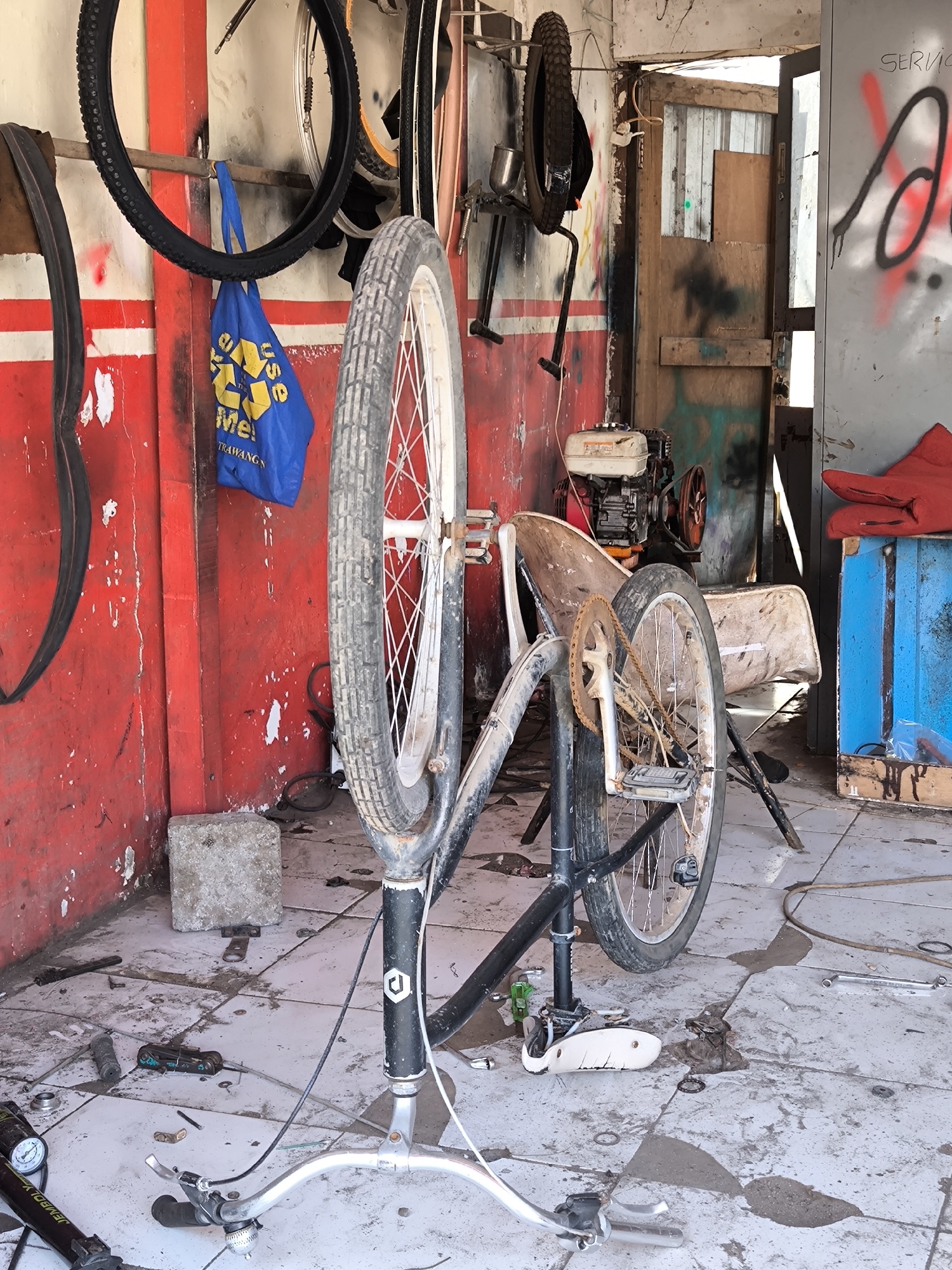 The image depicts the interior of what appears to be a bicycle repair shop. In the foreground, a bicycle is disassembled and propped up in a repair stand, with its front wheel removed. The front wheel is standing next to the frame, and the handlebars are lying on the floor. The rear wheel is still attached to the frame. A scattering of tools and bike parts are on the floor around the bike.
The walls of the shop are painted in alternating red and white stripes, with evidence of wear and staining. Several tires and a blue bag are hanging on the wall. To the right, an open doorway leads to an area with a chair and an engine. The back wall has graffiti. The floor is tiled, but the tiles are dirty and stained.
There are no people visible in the image.
The location is listed as Unknown, Indonesia.