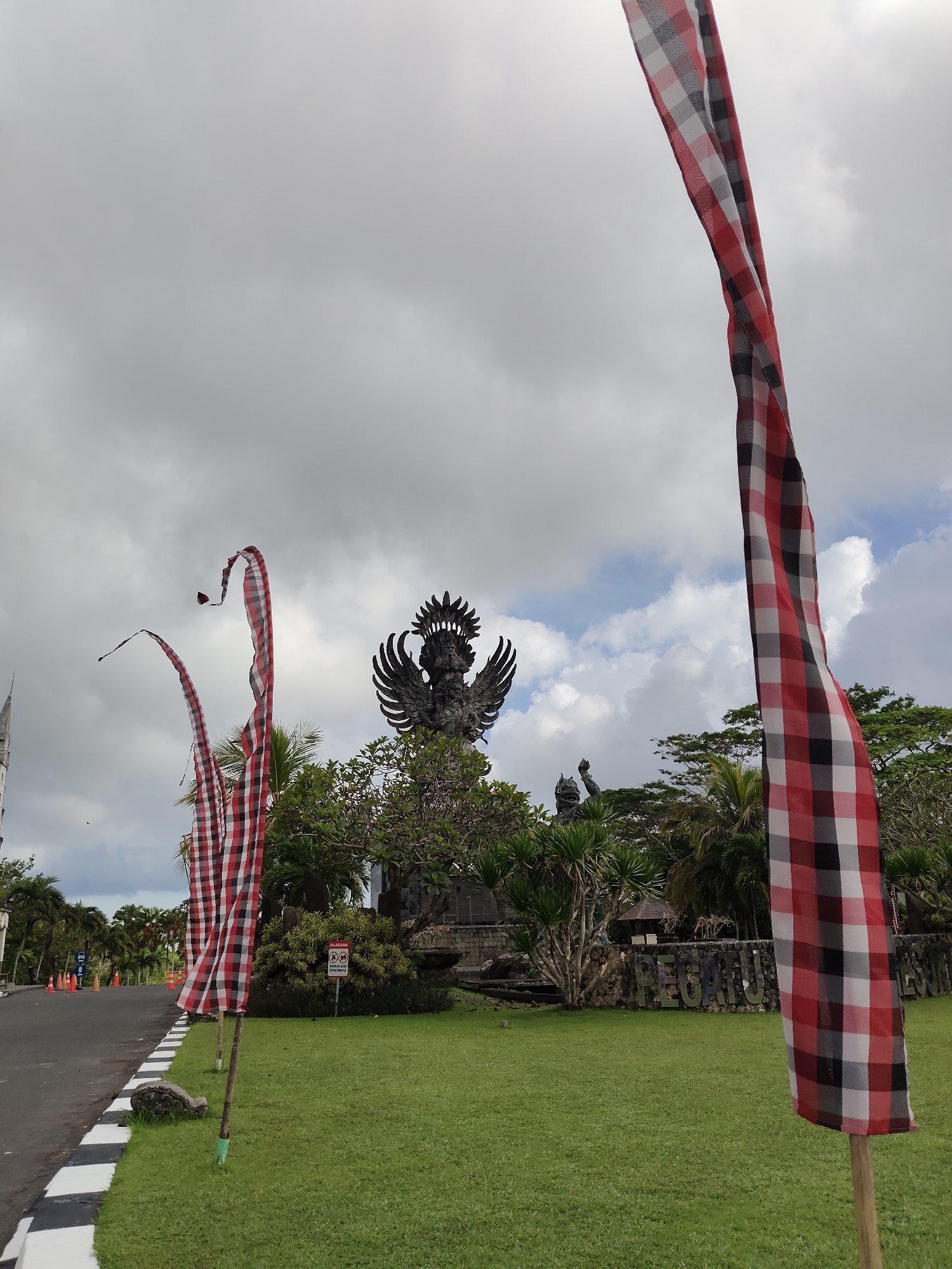 The image shows a view in Ungasan, Indonesia, featuring a large statue in the distance. The statue appears to be a figure with wings, standing above trees and greenery. In the foreground, red and white checkered banners are visible on poles. To the left, a road with black and white striped curbs is present. The sky is cloudy.