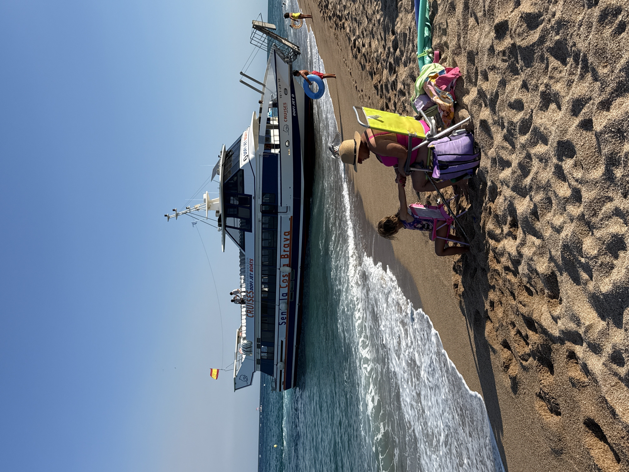 This image depicts a bright, sunny day at a sandy beach in Blanes, Spain. The foreground shows a woman and a child sitting on beach chairs facing a large white and blue passenger boat docked close to the shore. The woman, wearing a straw hat and a pink swimsuit, is holding the hand of the child who is looking down. Several beach towels and bags are scattered around the chairs, indicating a relaxed day at the beach.
The boat, labeled "CRUISES DOFI JET BOATS" and "Sen la Costa Brava," is a prominent feature of the scene, with several people visible on its deck. A boy is seen near the water's edge, holding onto a blue inflatable ring. Another person is further down the beach, near a ramp leading from the boat. The gentle waves of the Mediterranean Sea are lapping at the shore. The Spanish flag is flying on the boat.
The overall atmosphere is one of leisure and vacation. The clear blue sky and bright sunlight suggest it is midday. The sand is textured with footprints, indicating recent activity. The presence of the passenger boat suggests tourism and sightseeing are taking place in this coastal area.