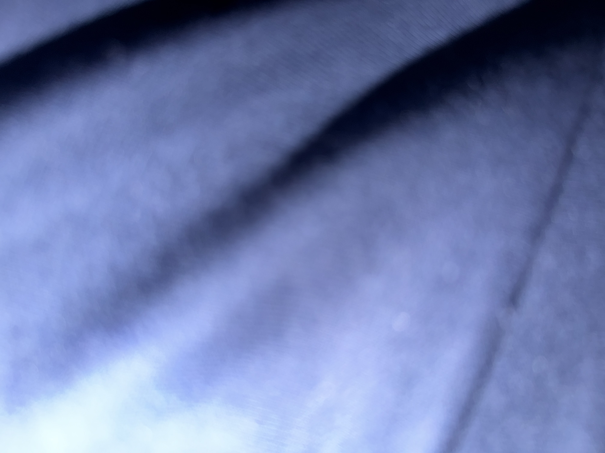 This image is an abstract and out-of-focus photograph of a textured, dark blue fabric. The lighting creates a sense of depth and highlights the folds and creases in the material, with a bright area on the left side fading into darker tones on the right. There are no discernible people, objects, or actions, and the environment is not identifiable. The image is not associated with Short Pump, United States of America.