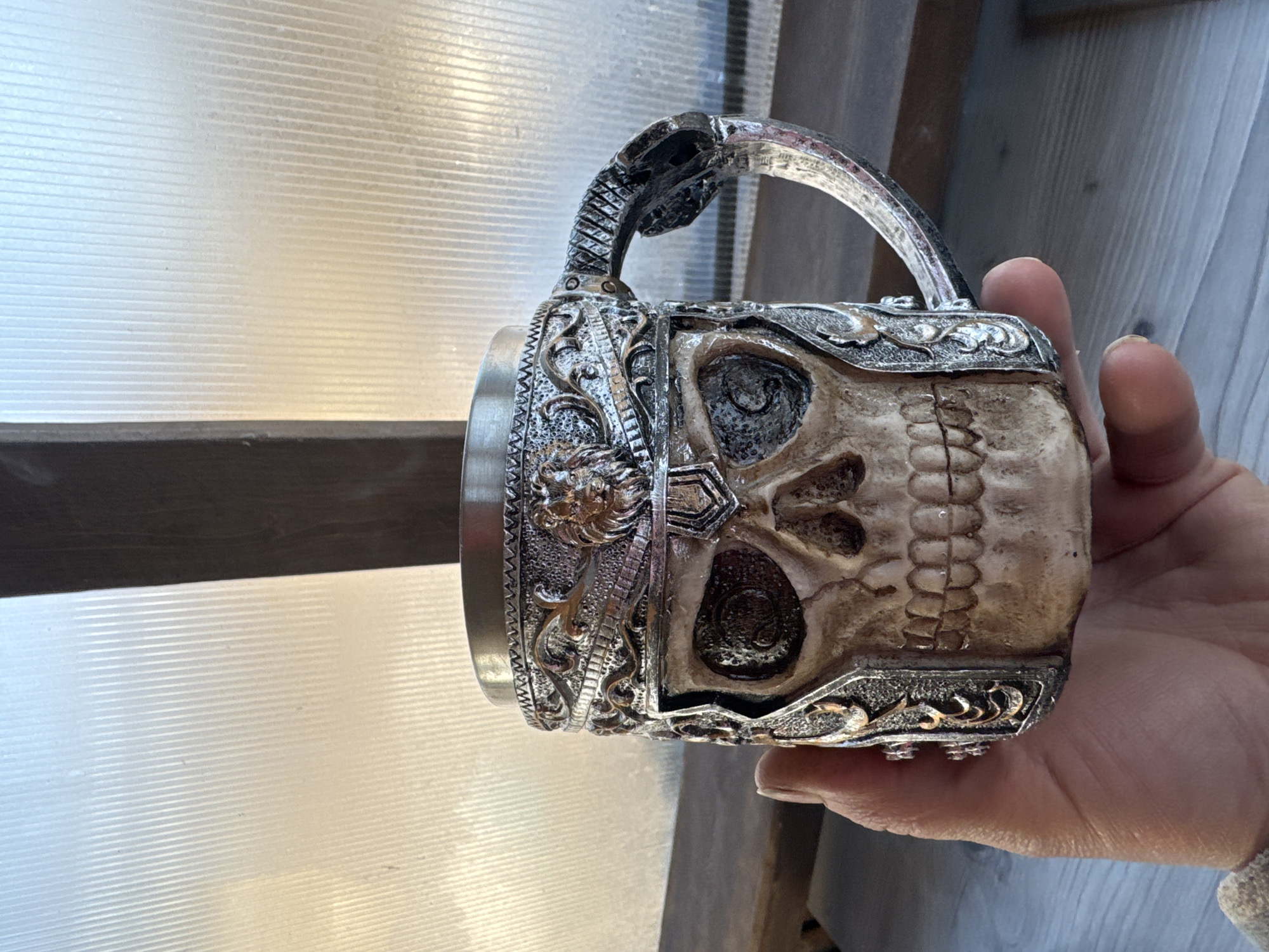 A decorative skull mug is held in a hand. The mug features a skull motif with ornate silver-colored metal detailing, including a handle shaped like a snake. The skull itself is a light beige color with darkened areas in the eye sockets and other crevices. The background includes a corrugated translucent panel above a dark wooden beam. The image was taken indoors, though the location is not evident from the visible elements. There is no indication the image was taken in Chaikovsky, Russia.