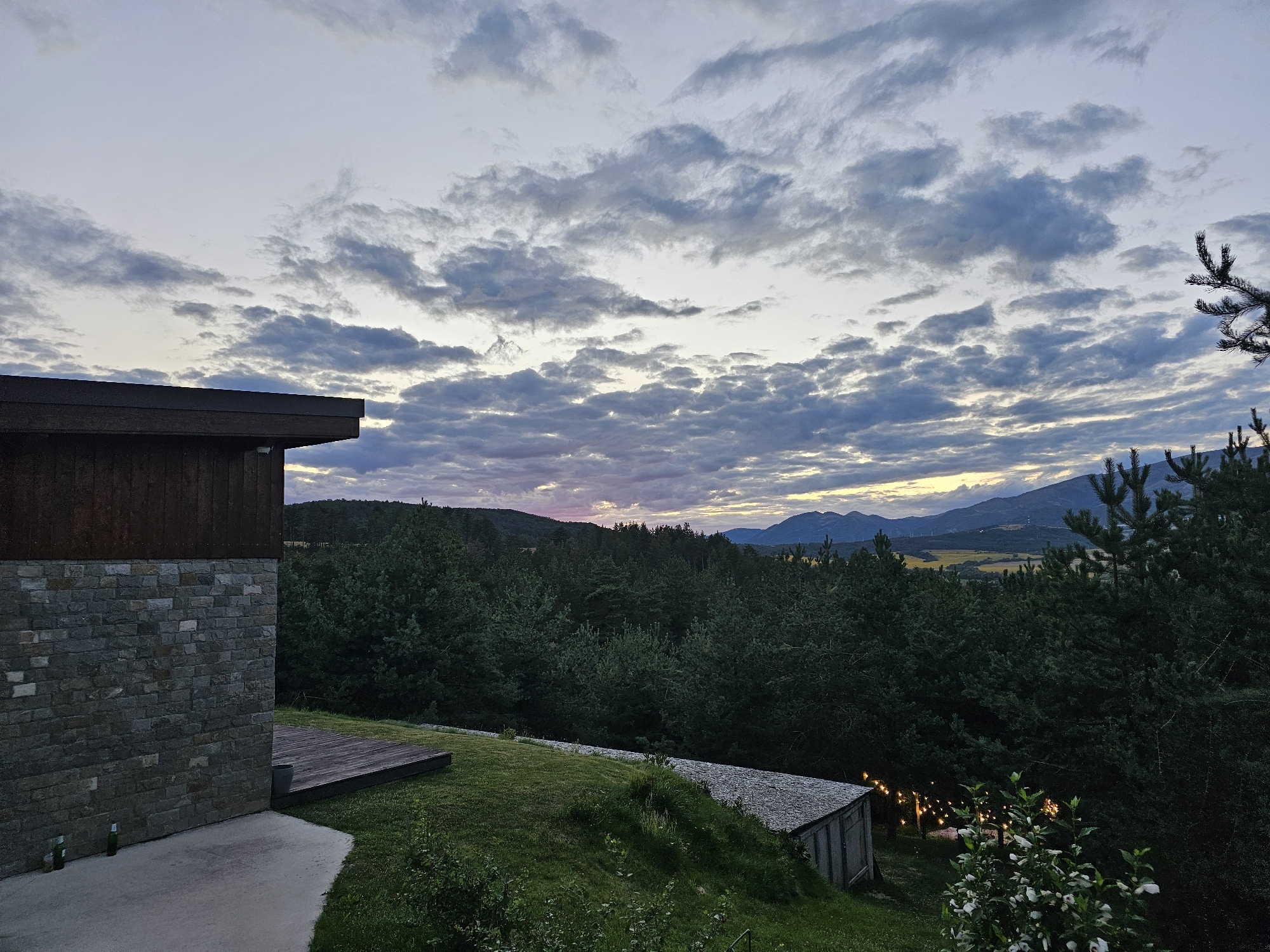 The image captures an outdoor scene at dusk, with a modern building in the foreground and a natural landscape in the background. The building features a dark wooden upper section and a stone-clad lower section, with a concrete patio and a wooden deck. Beyond the building, a dense forest stretches across the landscape, leading to rolling hills and mountains in the distance. The sky is filled with dramatic clouds, illuminated by the fading light of the setting sun, with hints of pink and yellow hues visible near the horizon. The overall atmosphere is serene and tranquil, suggesting a rural or semi-rural setting. There are no people or animals visible in the image. The lighting indicates it is either dawn or dusk.