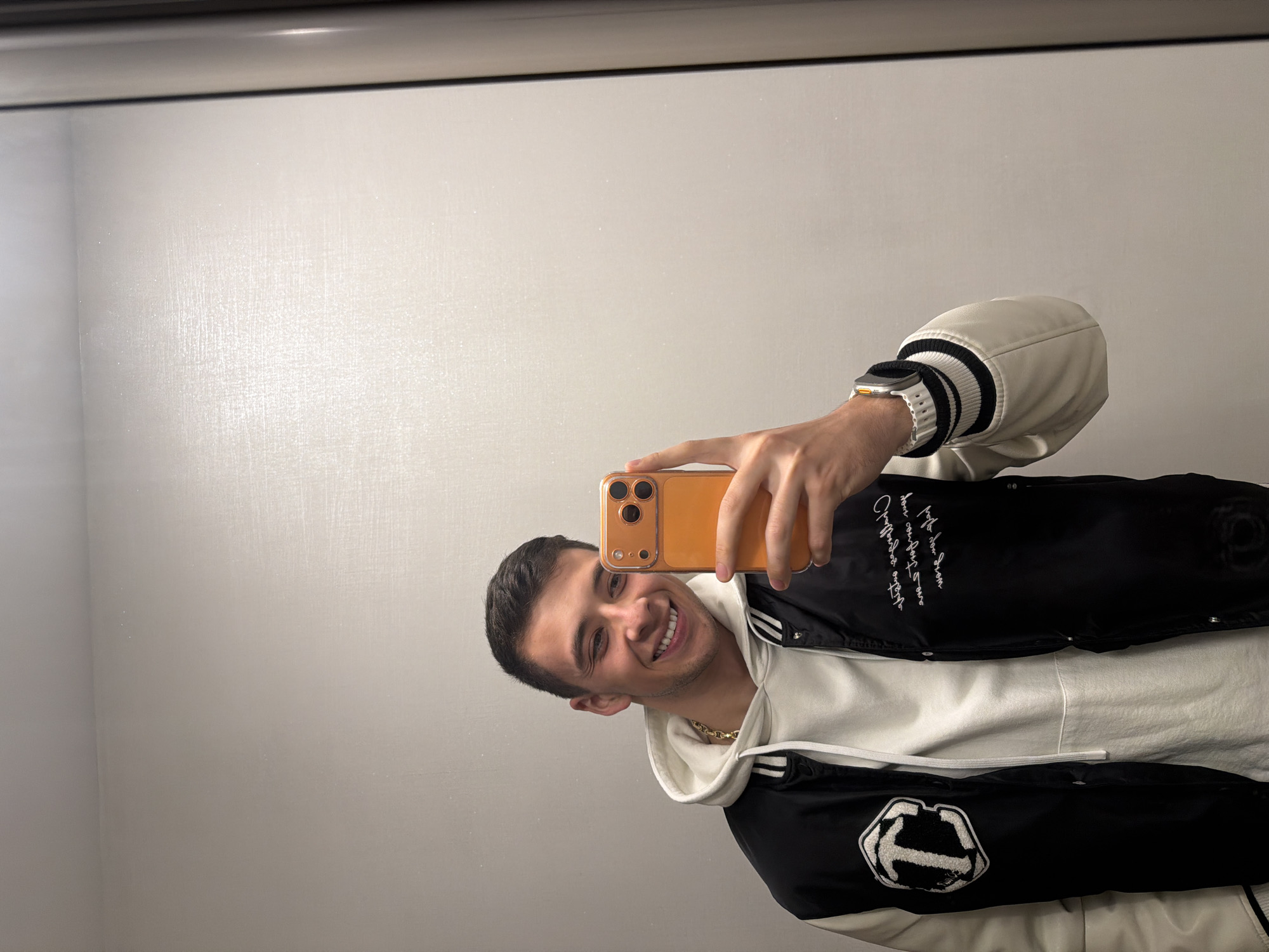 A young man is captured in a mirror selfie, holding an orange-cased smartphone with a triple camera system. He smiles broadly, displaying a cheerful and engaged expression. He is wearing a black varsity-style bomber jacket with white sleeves and black-and-white striped cuffs, layered over a white hooded sweatshirt. A gold chain necklace is visible around his neck. On his left wrist, he sports an Apple Watch Ultra with a white band. The background is a plain, light-colored wall, indicating an indoor setting reflected in a mirror. A dark, narrow frame or shelf runs horizontally across the top of the reflection.
Notable details on the jacket include white cursive writing on the upper left chest area, which is partially legible and appears to say "Classic Edition" or similar script. On the lower right side, a prominent white, textured patch resembling a stylized letter (possibly an "E" or "P") or an abstract logo with a fuzzy outline is present. The man's head is tilted slightly, and the image is bright, suggesting adequate indoor lighting.
