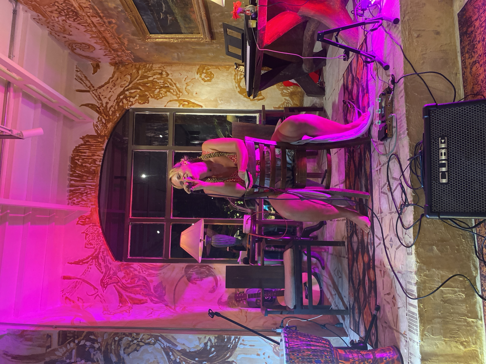 A blonde woman with braided hair is singing into a microphone while sitting on a chair on a stage. She is wearing a red and white floral dress and jewelry. The stage is decorated with murals and has musical instruments, lamps, and electronic equipment. The scene is lit with pink and red lights. The location is an indoor venue with windows looking out into the night.