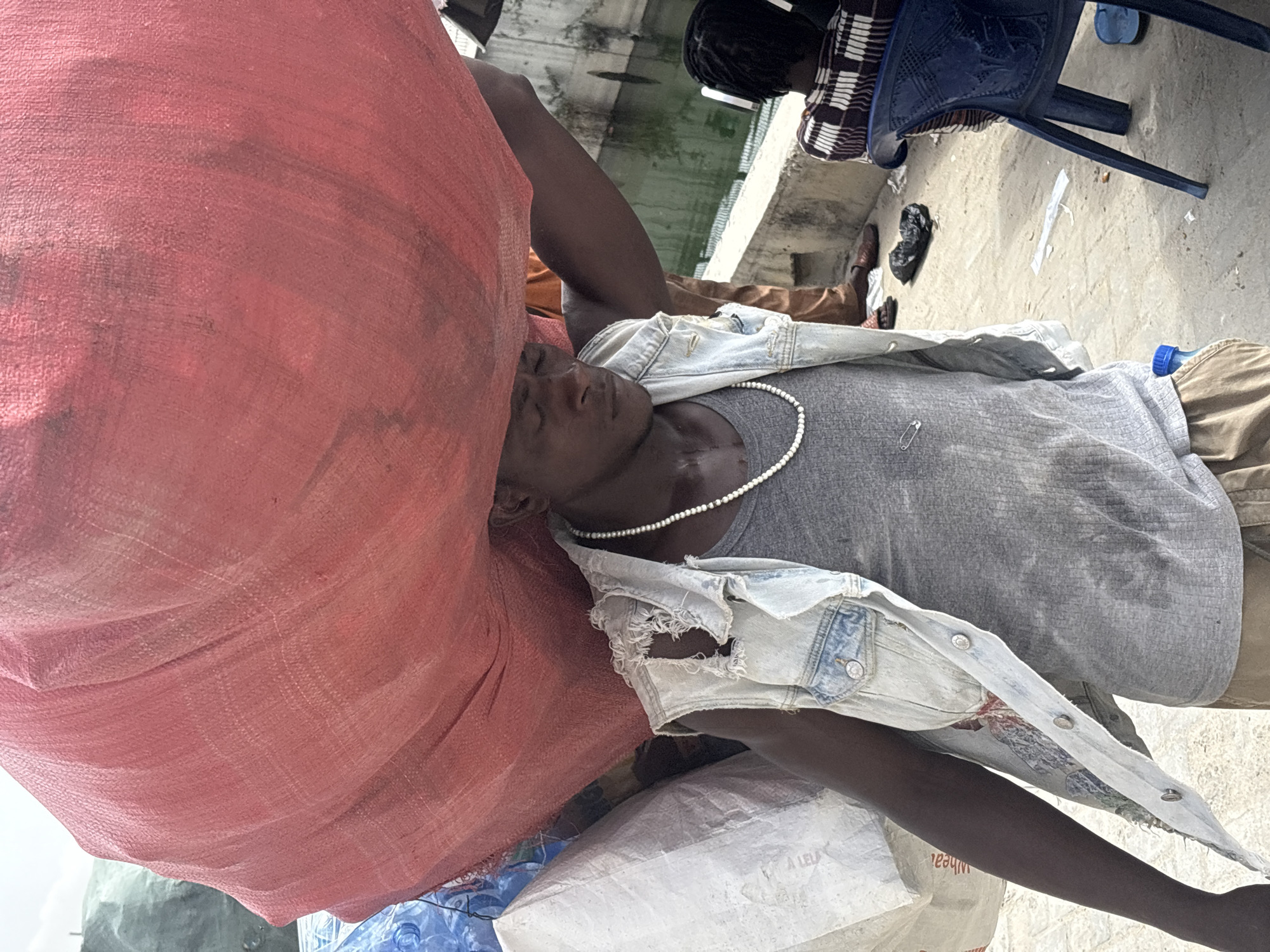 A person wearing a gray t-shirt, a denim jacket, and a pearl necklace lies under a large red sack. The scene is in Igboefon, Nigeria. Other details include: a blue chair, a person wearing a black and white checkered top, a water bottle, a white sack, and miscellaneous debris on the ground.