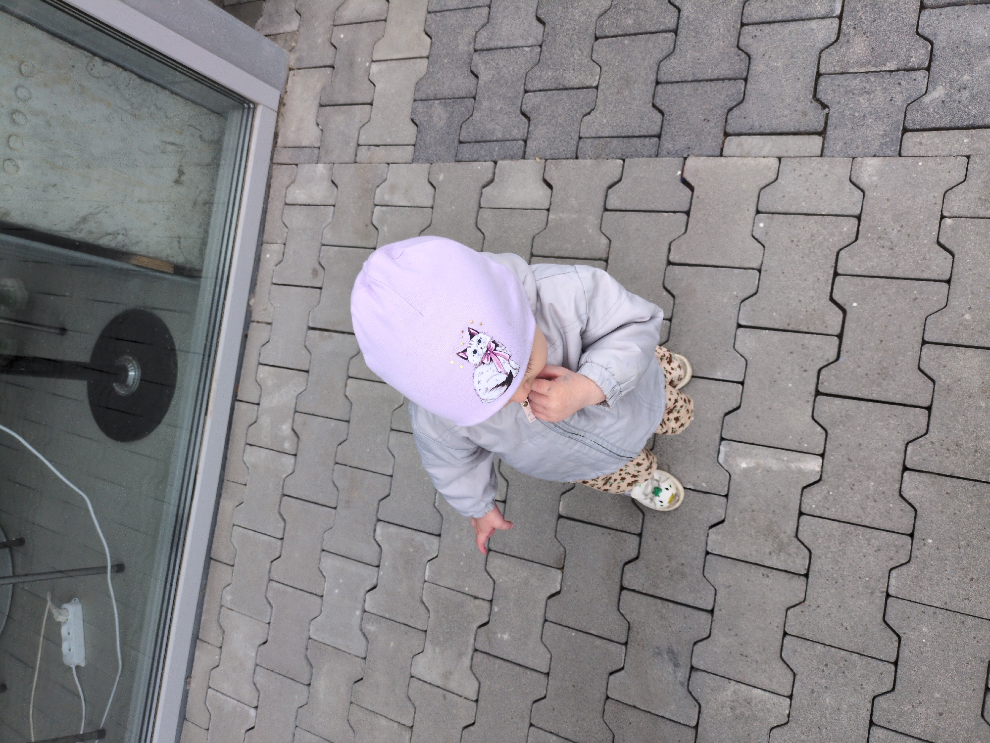 An overhead shot shows a toddler wearing a light grey jacket and a lavender beanie with a cat graphic on it, standing on interlocking gray pavers. The toddler's patterned leggings and white shoes are visible. The toddler is holding an object to their mouth with their right hand. The background features a reflective glass window with a light-colored wall and circular indentations visible behind it. A white electrical extension cord with a power strip is plugged in and visible on the floor inside. The scene appears to be outdoors in an urban environment.