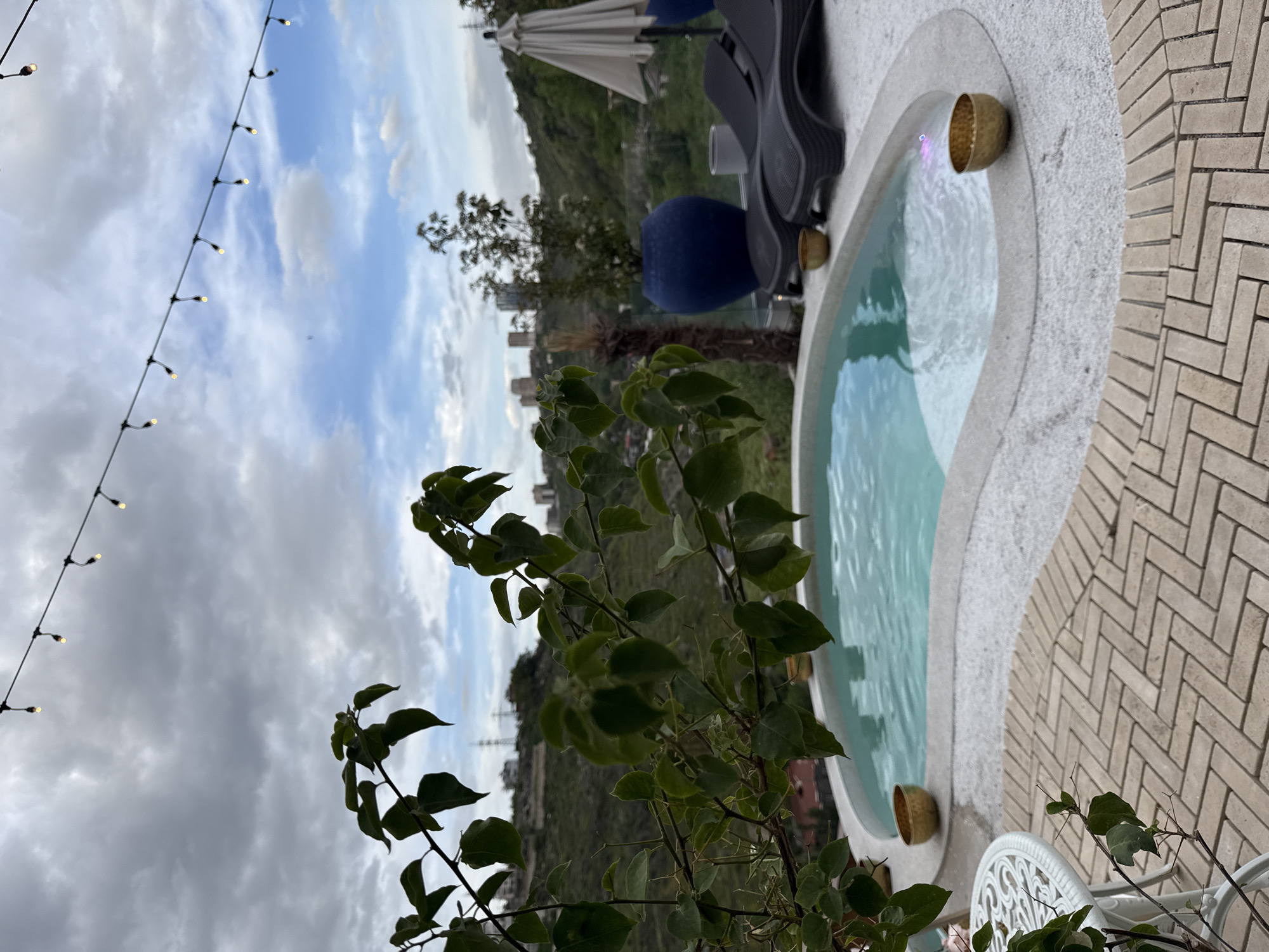 The image shows an outdoor scene, potentially a patio or terrace, in Yerevan, Armenia. A small, kidney-shaped pool or jacuzzi filled with water dominates the right side of the frame. Several metal containers are placed around the pool.
Behind the pool, in the background, there's a cityscape with buildings and greenery suggesting a hilly or mountainous terrain. In the mid-ground, there's outdoor furniture, including a sun umbrella, a dark-colored lounge chair, and a blue object, potentially a cushion or decor.
In the foreground, there's a plant with green leaves, partially obscuring the view, as well as a white table. String lights are visible in the sky. The area around the pool is paved with brick.
