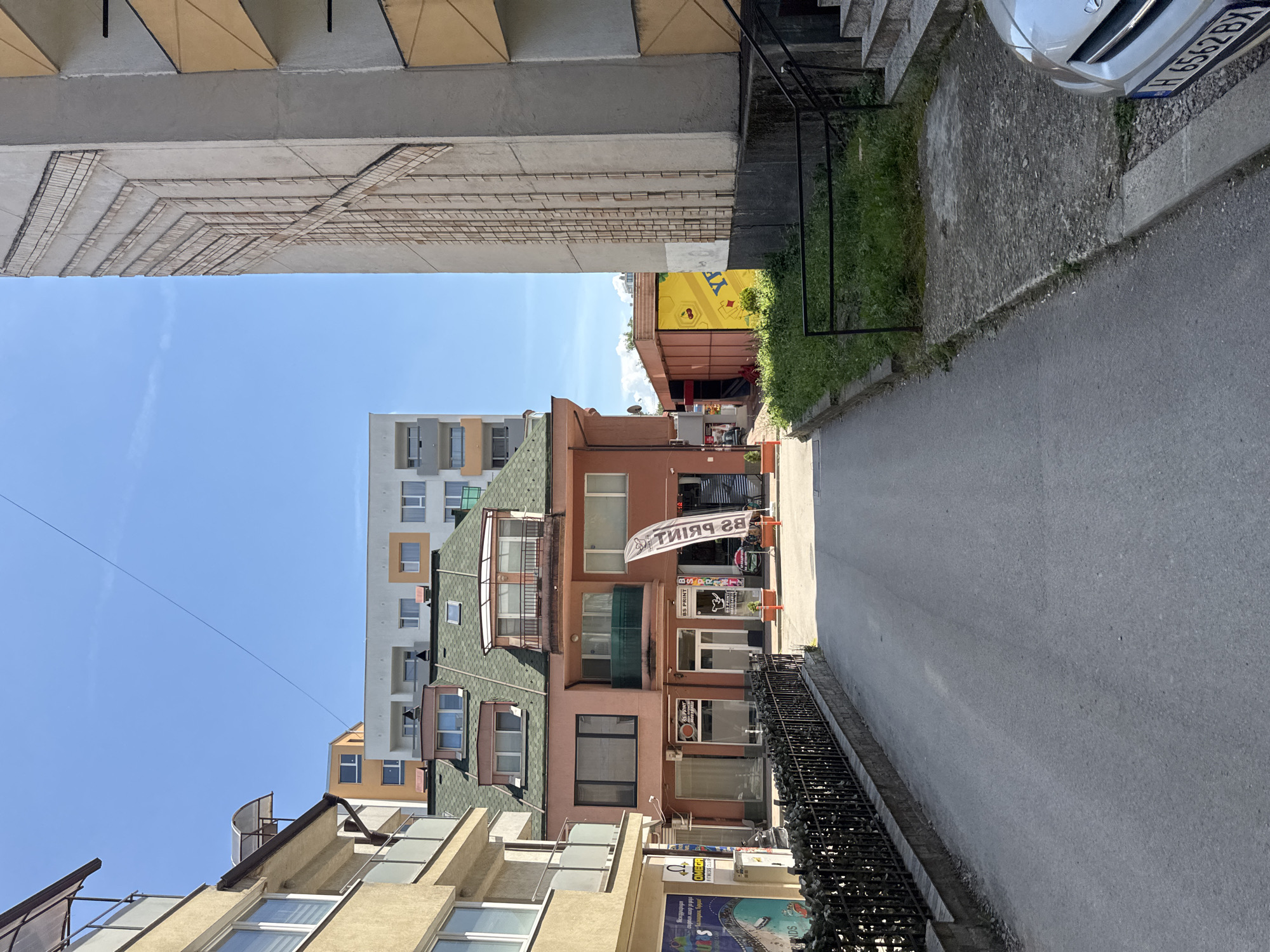 The image depicts a narrow street in Sofia, Bulgaria, lined with buildings of varying architectural styles. On the left, a light-colored building with balconies and modern windows stands tall. Adjacent to it, a multi-story building with a green, sloped roof occupies the foreground, featuring windows and a balcony. Below this structure, commercial spaces with glass storefronts are visible. A black metal fence runs along the left side of the street.
On the right side of the street, a tall, grey building dominates the view, characterized by a distinct vertical pattern of indentations and protrusions. Further down the street, a vibrant yellow structure with a red frame and a graphic of cherries is partially visible. The street is paved and appears to be an alleyway or a service road. A portion of a silver car with a license plate starting with "H 6562" is parked on the right side of the street near the bottom right corner of the image. The sky is clear and blue, indicating a sunny day. The overall scene suggests an urban environment with a mix of residential and commercial properties.