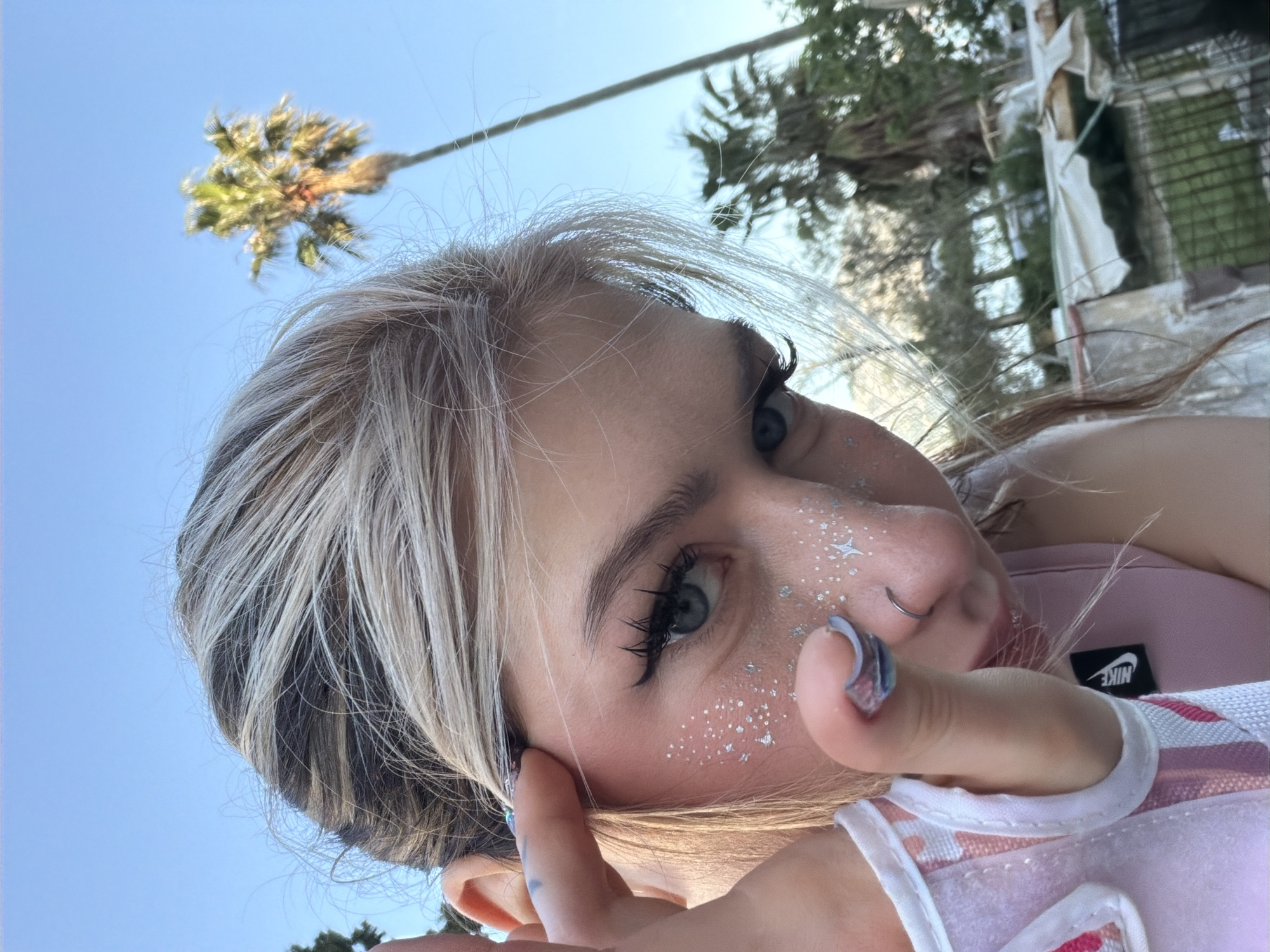 This close-up, low-angle portrait features a young person, likely female, set outdoors against a clear blue sky in Budva, Montenegro. The subject has fair skin, blue eyes, and streaky blonde/ash-blonde hair with visible darker roots. They wear prominent eyelashes, possibly false, and have a silver nose ring on their left nostril. Silver glitter and small star-shaped appliqués are scattered across their cheekbone and the bridge of their nose, suggesting a festive or playful aesthetic.
The person's hand, adorned with blue, white, and purple patterned nail polish, is raised near their face, either touching their hair or posing for the shot. The image is taken from a tilted perspective, rotating the subject's head. The background consists of a bright, clear blue sky with a distinct palm tree silhouetted against it. Further back, additional palm trees and blurry structures with white and green elements can be seen, indicating an outdoor, possibly urban or resort setting.
A section of a pink or light-colored garment, possibly a tank top or backpack strap, is visible in the lower right corner. A black rectangular label with the white text "NIKE" is clearly visible on this material. The photograph captures a casual, bright moment under natural daylight, conveying a relaxed and perhaps whimsical mood.