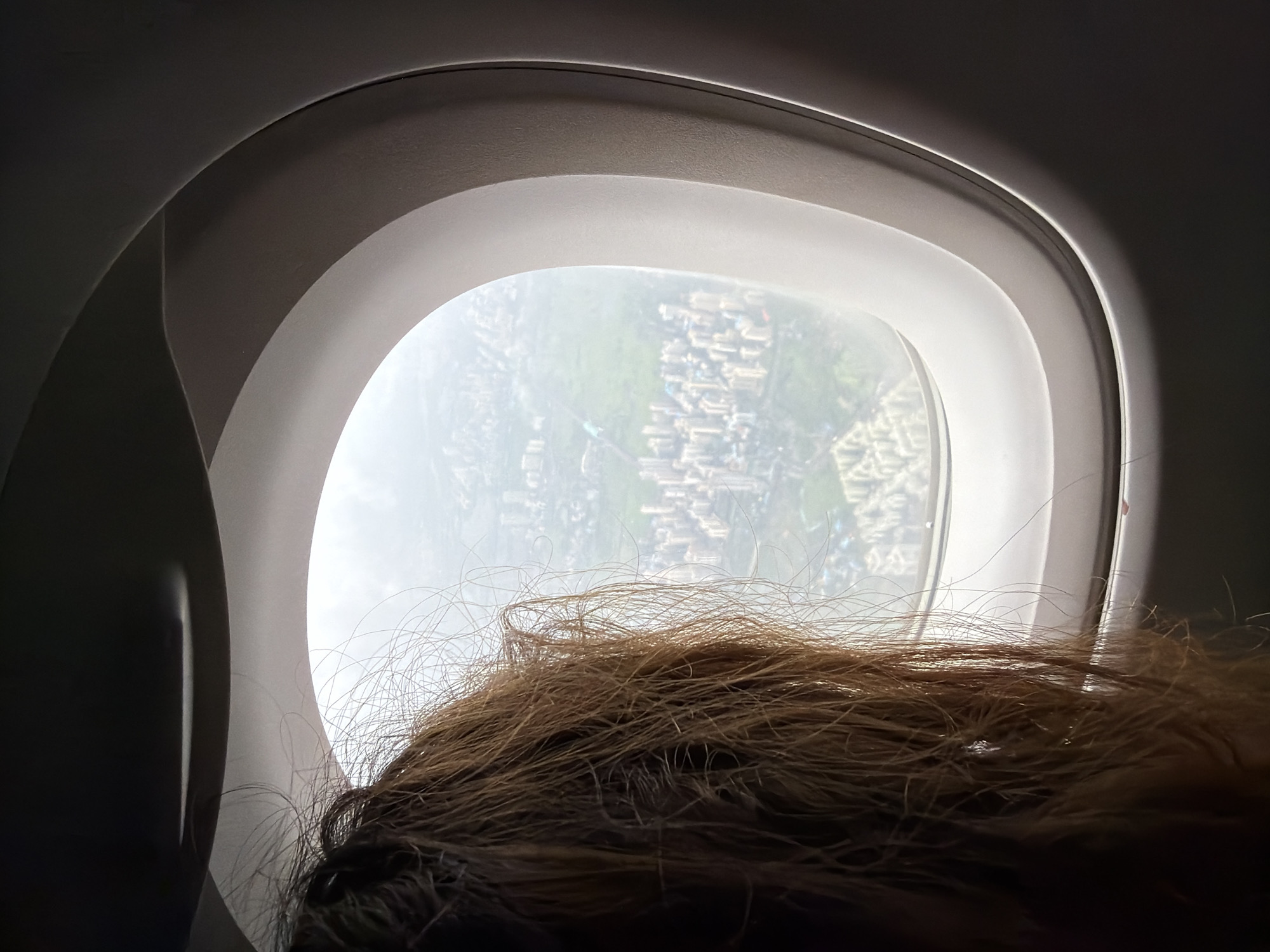 The image is a close-up, over-the-shoulder view of a person's head with brown hair, looking out of an airplane window at the city of Panvel, India. The airplane is in flight, and the view outside shows a dense urban area with numerous buildings, some green spaces, and what appears to be a hazy sky. The airplane window frame is prominently visible, with its characteristic oval shape and layered design. The lighting suggests it is daytime. The focus is on the contrast between the dark interior of the cabin and the brighter, distant cityscape.