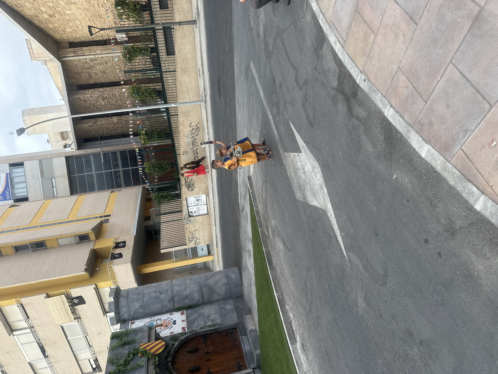 The image depicts a street scene in Alicante, Spain. In the foreground, two young boys are walking on a street, one holding a toy sword. They are wearing similar yellow and blue outfits. The street has a white arrow painted on it, indicating a direction. To the left of the street is a decorative facade resembling a castle gate with ivy growing on it. Behind the castle facade, a multi-story building with yellow and beige tones is visible, adorned with black flags featuring a cross. Further in the background, there is another building with a stone facade, and string lights are hung across the fence in front of it. The sky is overcast. The general setting suggests an outdoor urban environment, potentially a public space or a street during an event or festival.