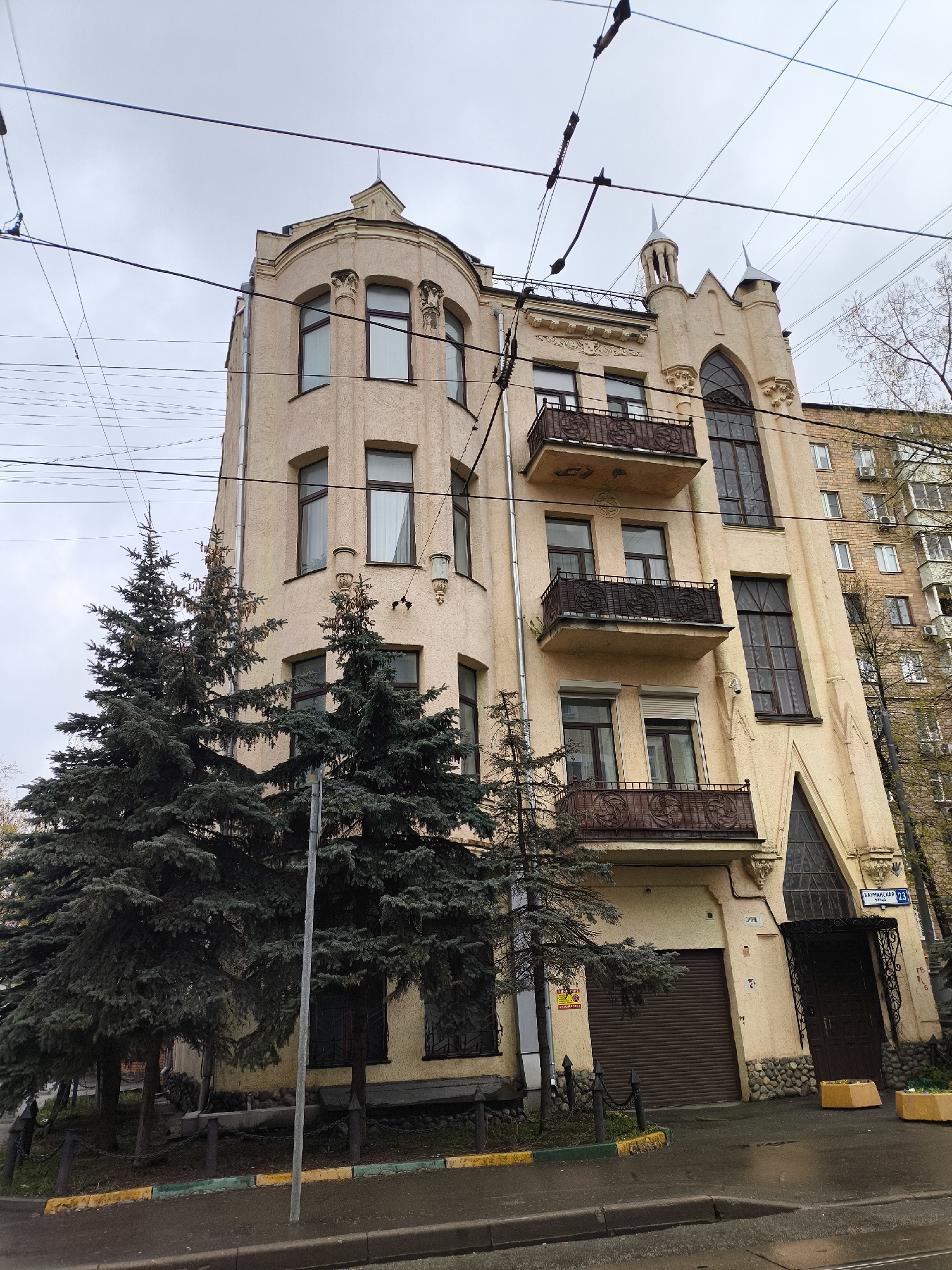 The image presents an ornate, multi-story building on an overcast day in an urban setting, identified as Sadovaya-Kudrinskaya 23 in Moscow, Russia. The building, rendered in a pale yellow or light beige, showcases a distinctive architectural style, possibly Art Nouveau or Gothic Revival, with intricate details.
Its most prominent feature is a rounded corner section on the left, adorned with multiple dark-framed windows and decorative architectural elements, including carved corbels and a small turret-like structure at the top. The right side of the building features a striking tall, narrow section with a large, pointed Gothic-style arch window, topped by small spires. Several balconies with elaborate dark iron railings are visible on this facade.
The ground floor includes a dark brown roll-down garage door and a matching main entrance door, both accented by a base of rough stones. Large, dark green evergreen trees (likely spruce or fir) partially obscure the lower left portion of the building, providing a natural element against the constructed environment.
Overhead, numerous utility lines crisscross the sky. The street in the foreground is visibly wet, suggesting recent rain, and features a curb painted with sections of yellow and green. A street lamp stands near the trees. In the background to the right, a plainer, multi-story brick residential building is visible.
Visible text includes the blue street sign "САДОВАЯ-КУДРИНСКАЯ 23" (Sadovaya-Kudrinskaya 23), confirming the location. Other signs on the ground floor indicate "РЕМОНТ ОБУВИ" (shoe repair) and "АВТОШКОЛА" (driving school). No people are present, and the scene is static, capturing the building's unique facade and its surroundings under a cloudy sky.