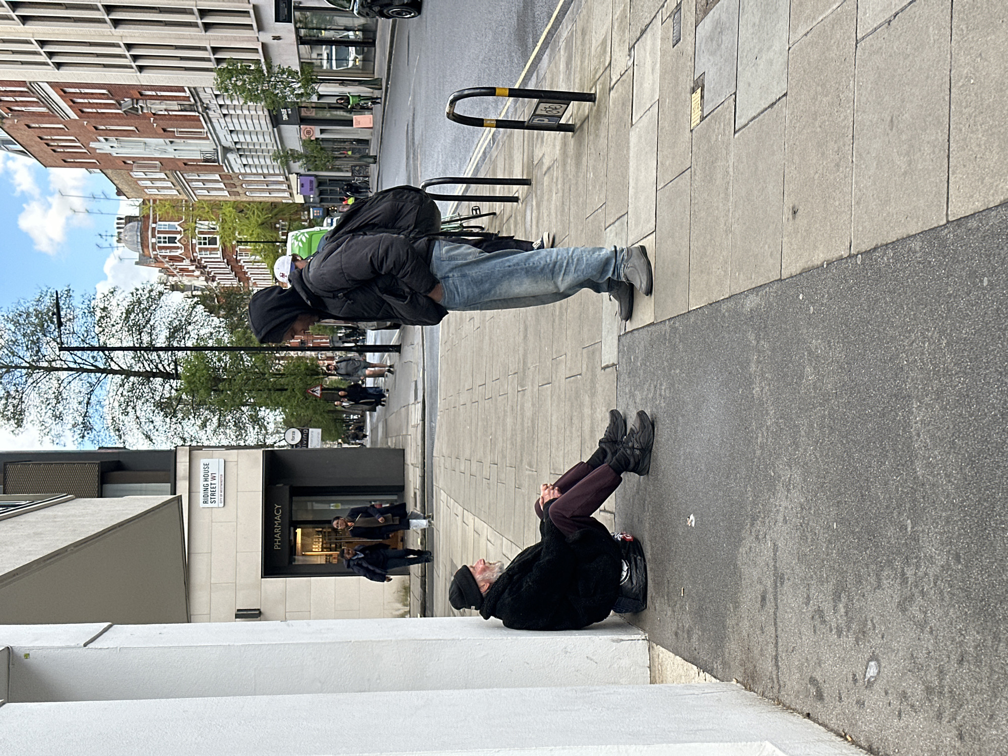 Two individuals are engaged in a physical balancing act on a city street. One person, wearing a black jacket, blue jeans, and gray shoes, is in a horizontal plank position, supporting their weight on their hands and feet. The second person, an older individual with a gray beard and wearing a black beanie and dark clothing, is positioned at one end, seemingly providing counter-balance or support. The scene is set on a sidewalk, with a building on one side and a street with vehicles and pedestrians in the background. A street sign indicates the location as Riding House Street W1, City of Westminster, London. A pharmacy is visible nearby.