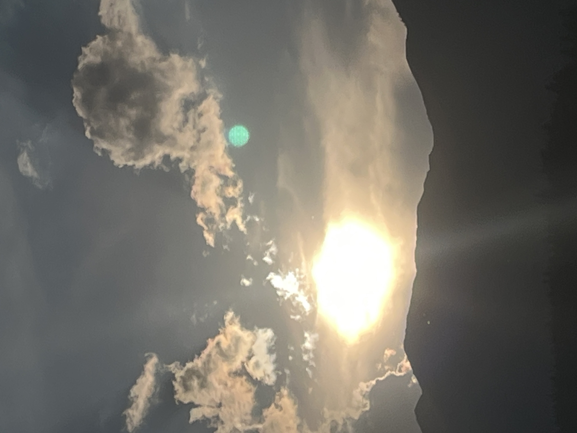 This media file depicts a sky filled with dramatic clouds and the bright sun, likely captured during a scenic view in Garmisch-Partenkirchen, Germany. The sun is a dominant element, appearing as a very bright, almost square-shaped source of light in the center-right of the image. Sunbeams radiate outwards, illuminating the surrounding clouds.
The clouds are varied in texture and color. Some are dark and imposing, appearing almost silhouetted against the brighter parts of the sky, particularly on the far right edge which suggests a very dark, possibly shadowed, area. Other clouds are illuminated by the sun, appearing bright white and golden, with wispy edges. There's a notable green circular glint in the sky, possibly a lens flare or reflection.
The overall scene conveys a powerful natural display of light and atmosphere. The composition, with the stark contrast between light and shadow, and the dramatic cloud formations, suggests a dynamic and possibly turbulent weather condition, or simply a striking sunset or sunrise. The image evokes a sense of awe and the grandeur of nature. No people, objects, or visible text are present in the frame. The time of day is likely either sunrise or sunset due to the intense sun and dramatic lighting.