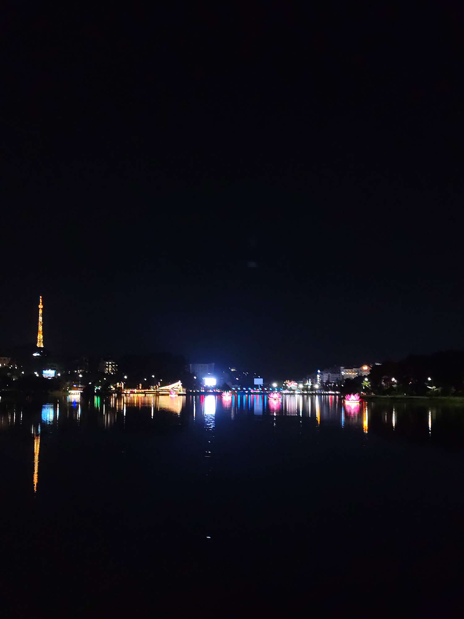 The photograph depicts a nighttime scene of Ho Xuan Huong Lake in Da Lat, Vietnam. In the background, a brightly lit communication tower is visible along with other city lights outlining the shoreline. A bridge adorned with colorful lights stretches across the lake, its reflection mirroring in the dark water. Floating lanterns, also illuminated, are scattered across the lake's surface. The water is calm, reflecting the various lights of the city and decorations. The sky is dark, with minimal visible features.