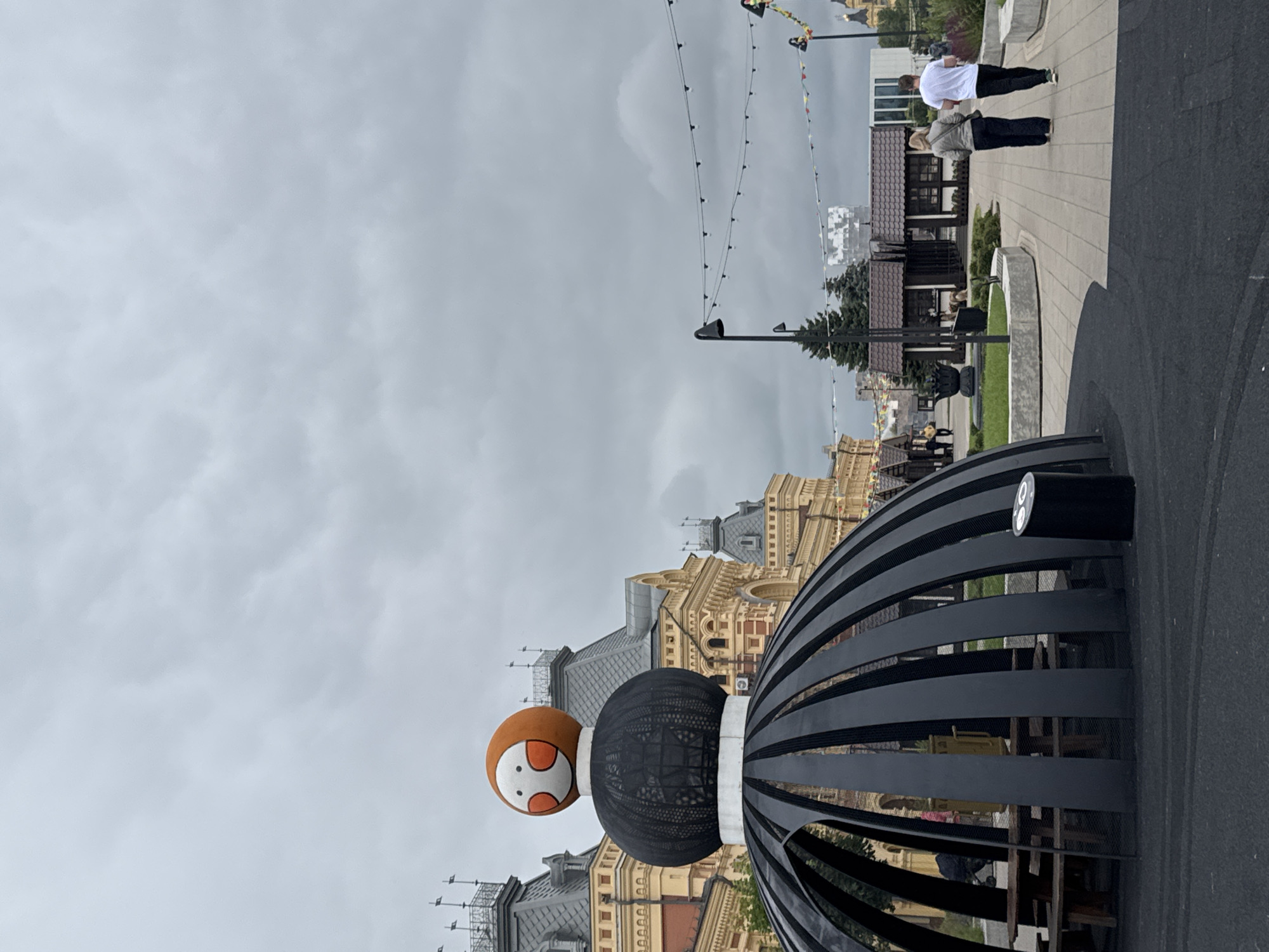 The image depicts a street scene in Nizhny Novgorod, Russia, featuring an abstract sculpture. The sculpture is large and dome-shaped, constructed of black metal ribs. Atop the dome sits a sphere with a cartoonish face, featuring orange cheeks and eyes. Behind the sculpture, the iconic architecture of the Nizhny Novgorod Kremlin is visible, characterized by its historic buildings with ornate details and grey tiled roofs. The sky is overcast, suggesting a cloudy day. In the foreground, a paved walkway leads past the sculpture and towards a line of small, modern buildings, possibly shops or food stalls, also with tiled roofs. Two individuals, a man and a woman, are walking away from the camera on the walkway. The man is wearing a white t-shirt and black pants, while the woman is in a grey jacket and black pants. The overall atmosphere is calm and everyday, with the sculpture adding a touch of artistic flair to the urban landscape.