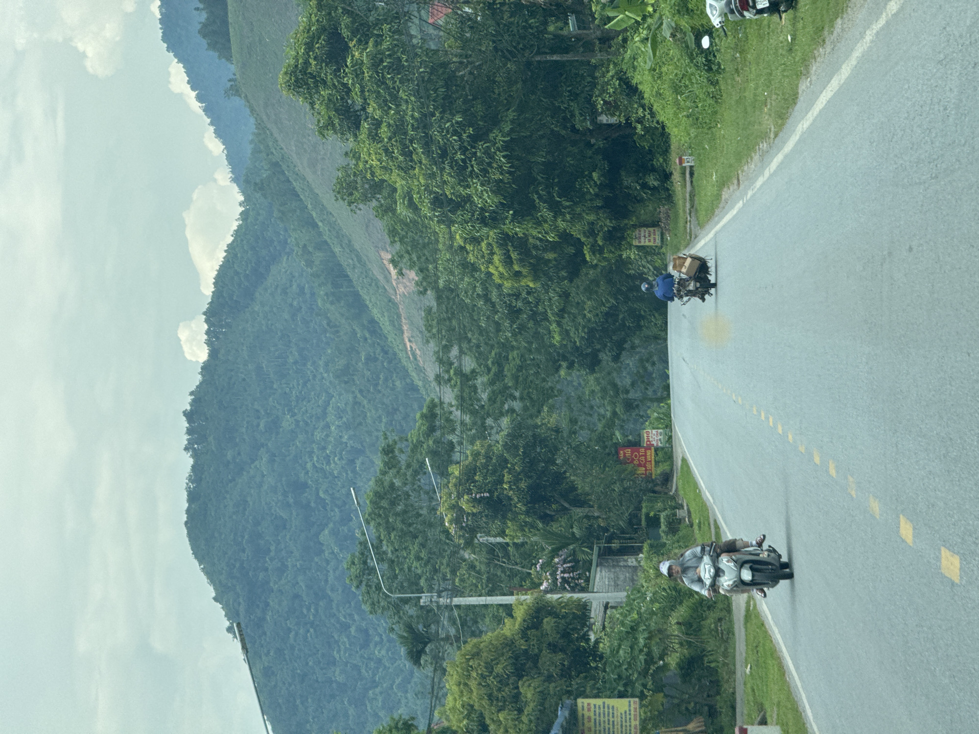 The image shows a paved road cutting through a landscape of lush green mountains under a cloudy sky. On the road, two individuals are riding motorbikes in the same direction. The motorbike in the foreground carries two people and the one behind appears to be carrying some cargo. The road has yellow dotted lines marking the center. On the side of the road are trees, vegetation, and utility poles. Based on the road signage, foliage, and mountainous terrain, the location appears to be along a road in the city of Hà Giang 2 Ward, Vietnam.