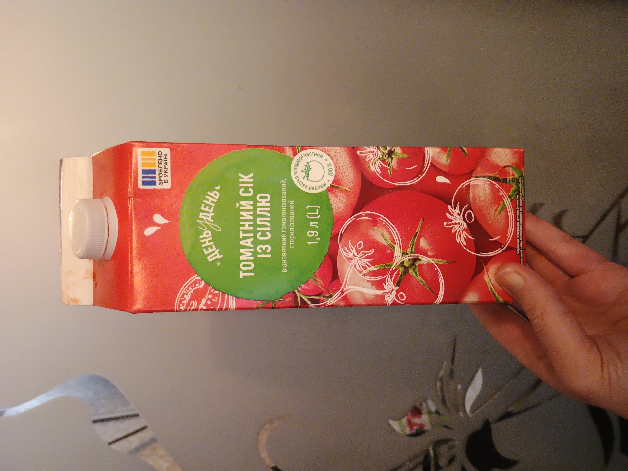 A hand is holding a carton of tomato juice. The carton is red and features a green circle with white text that says "ДЕНЬ" and "ДЕНЬ" in a stylized font. Below that, it says "ТОМАТНИЙ СІК ІЗ СІЛЛЮ" and "відновлений гомогенізований, стерилізований". Underneath that, it states "1,9 Л (L)". There is a small circular emblem on the right side of the carton with the text "частика плодовоі частини 100%" and an illustration of a tomato plant. A smaller logo at the top right of the carton reads "ЗРОБЛЕНО В УКРАЇНІ". The background is a wall with a decorative pattern. There is no information in the image that would allow for determining the location in the city of Zaporizhzhia, Ukraine.