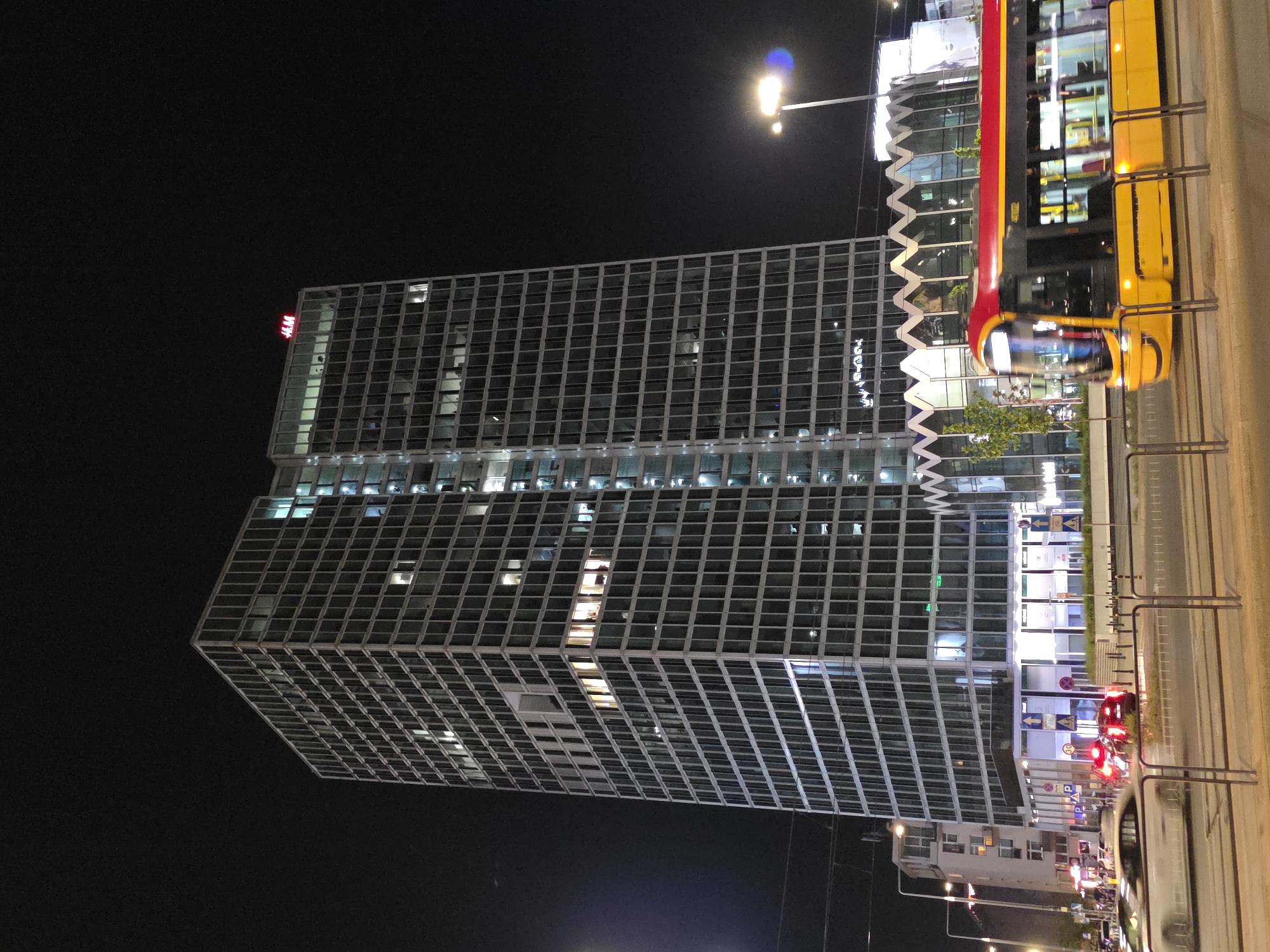 At night in Warsaw, Poland, a tall, square skyscraper with a grid-like facade dominates the scene. A red "HM" logo is visible at the top corner of the building. A yellow and red tram passes by on the right side of the frame. Below, cars with their red taillights are visible. Streetlights illuminate the area.