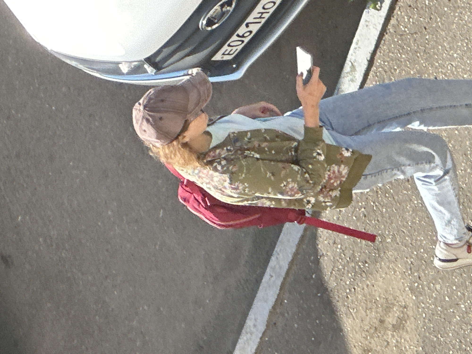 A person wearing a floral jacket, light blue t-shirt, and light blue jeans is walking past a car. The person is also wearing a brown baseball cap and carrying a red backpack. They are holding a white smartphone in their right hand. The scene takes place outdoors on a paved surface next to a car with a license plate reading "E061HO". This image was captured in Podolsk, Russia.
