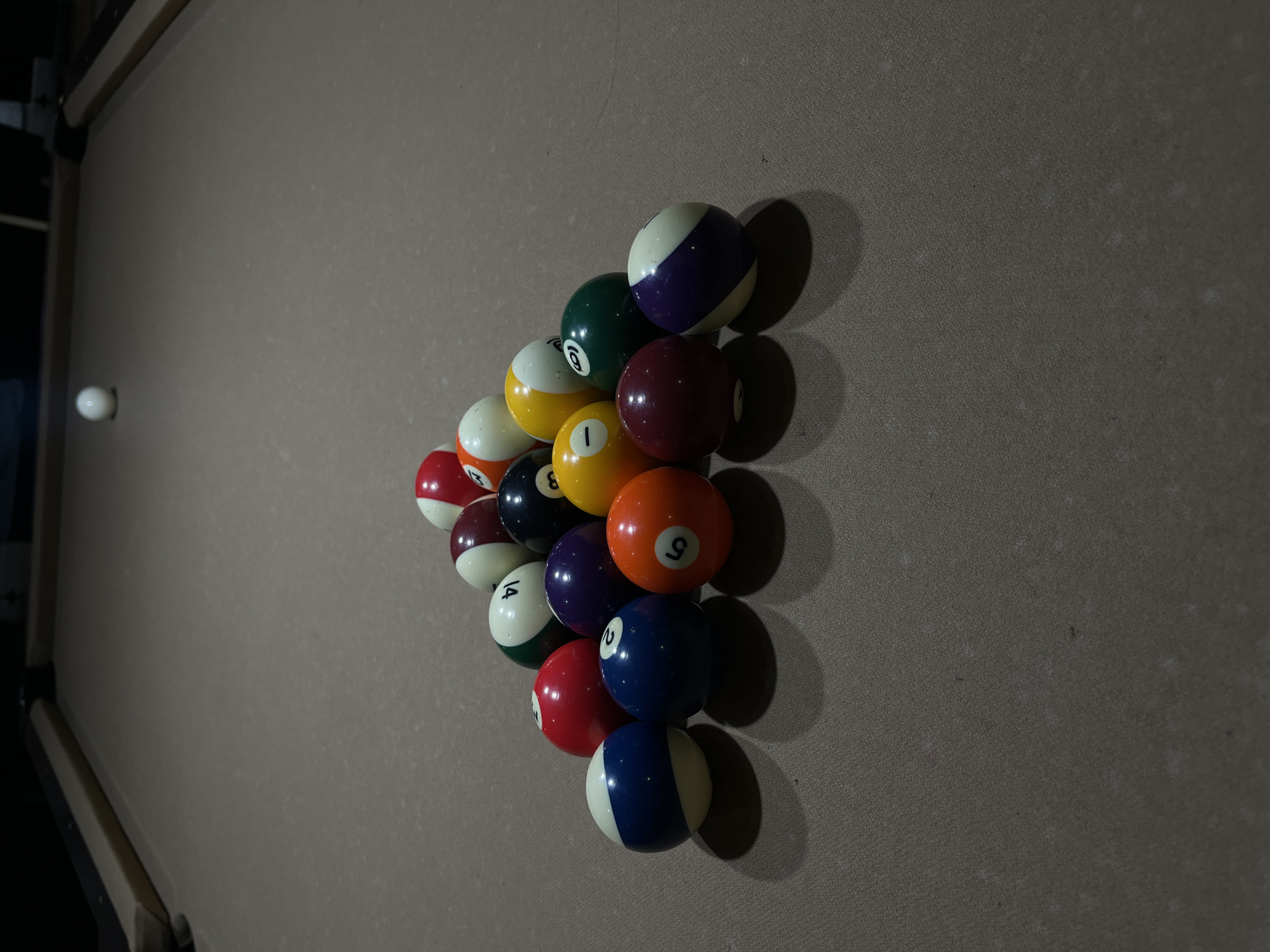 A still life photograph taken indoors depicts a billiard table with a racked set of pool balls. The pool balls are arranged in a triangular formation towards the right side of the frame, comprising solids and stripes. A single, white cue ball sits on the left side of the table. The table is covered in a gray-brown cloth. The pool table has a wooden rail along its perimeter. The lighting is somewhat subdued and casts shadows from the balls onto the table. The exact location is not specified.