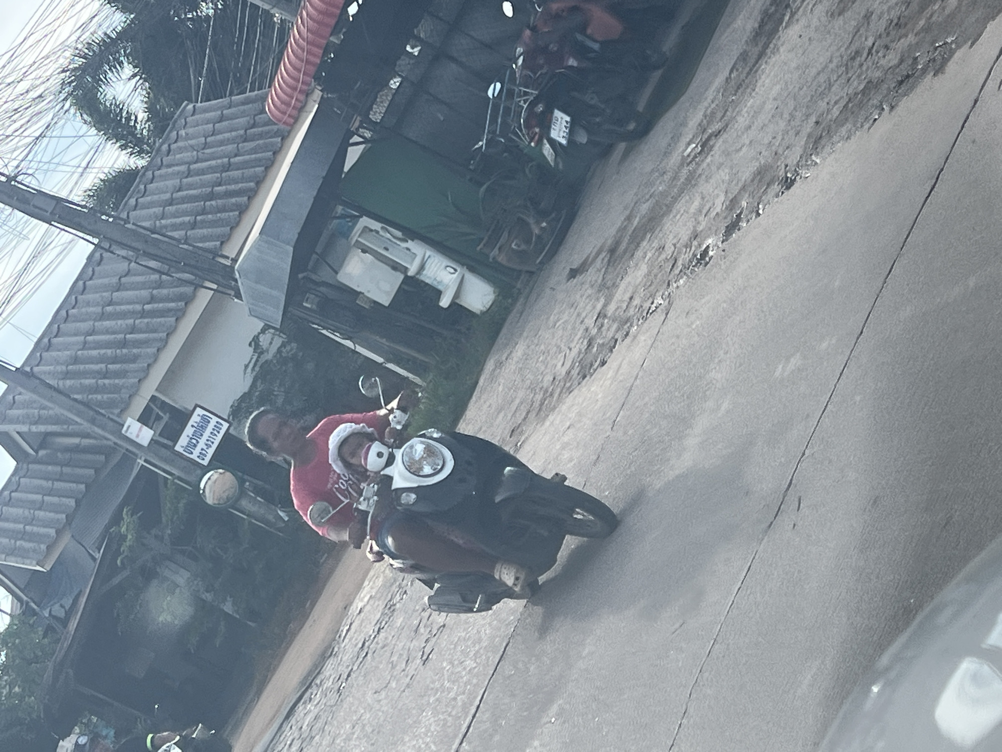 A tilted outdoor shot shows an individual riding a scooter on a road in บ้านบางรักษ์, Thailand. The individual, wearing a red shirt and Santa hat, rides a dark-colored scooter. A smaller individual in a Santa hat is seated in front of the rider. The road surface is paved. In the background, a building with a gray tiled roof is partially visible. Two motorcycles are parked near the building. Utility wires are visible overhead. A sign with Thai script is also visible.