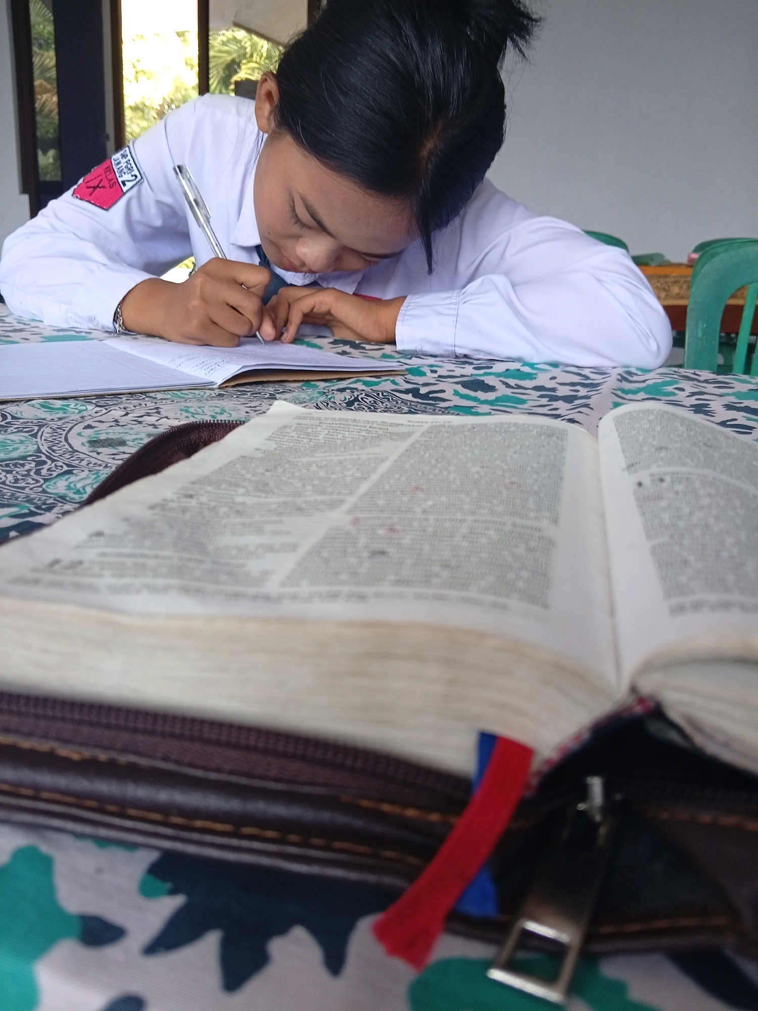 A young Indonesian female student in a white uniform with a red and white patch on her left sleeve is diligently writing in a notebook. She is seated at a table with a patterned tablecloth, and an open book lies in the foreground. The background shows a window with sunlight streaming in, suggesting an indoor setting, possibly a school or home in Lawang, Indonesia. The girl's focus is on her writing, indicating an academic or study-related activity.