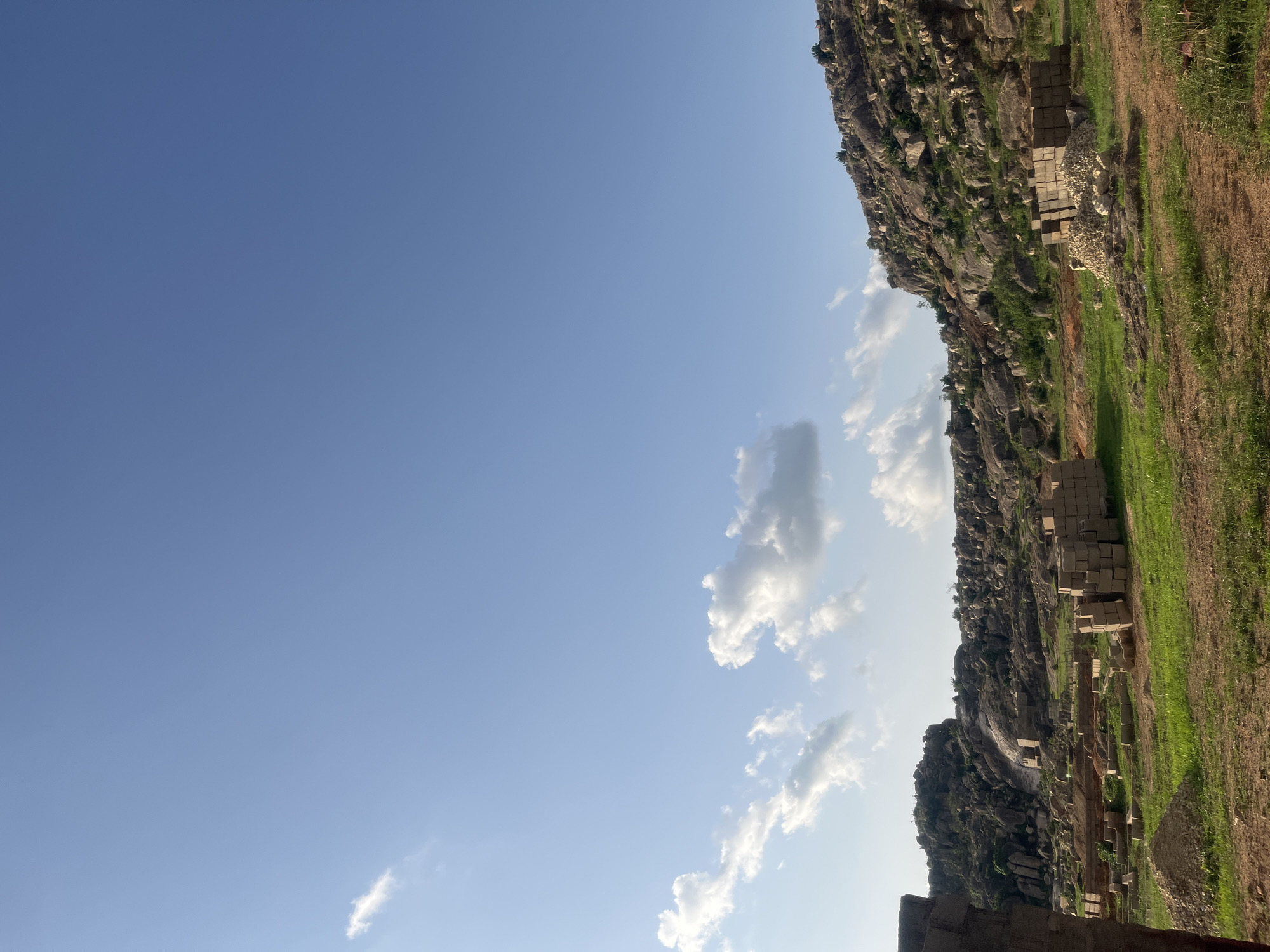 The image displays a rugged landscape in Gura Top, Nigeria, under a clear blue sky dotted with scattered white cumulus clouds. In the background, prominent rocky hills covered with numerous large boulders and sparse green vegetation dominate the scene. The mid-ground features uneven terrain with patches of green grass and brown soil, interspersed with multiple stacks of grey concrete blocks and piles of aggregate, indicating construction activity. A partially constructed wall is visible on the far left edge of the frame. No individuals are present.