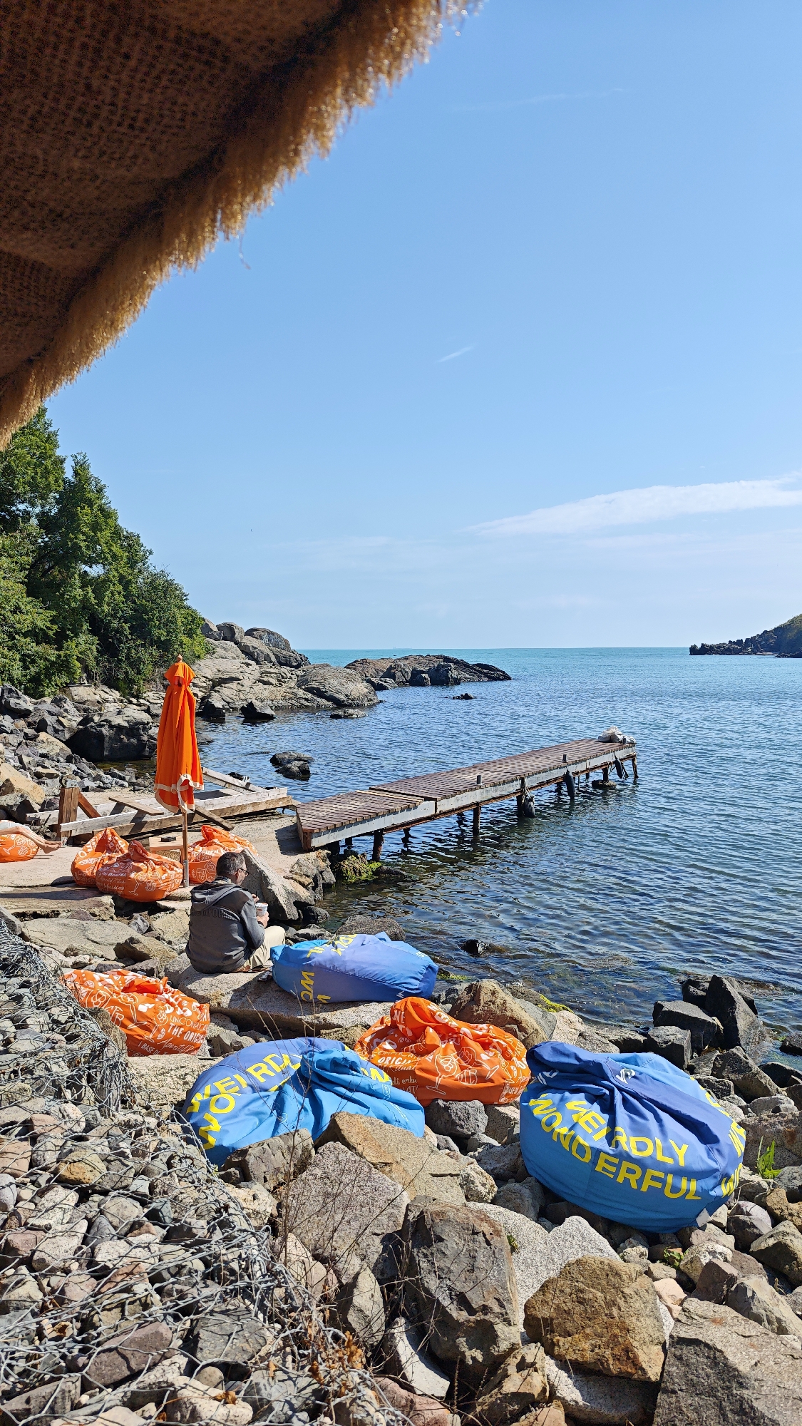 The image presents a serene coastal scene in Sozopol, Bulgaria, on a bright, sunny day. The vantage point is from beneath a thatched or woven canopy, which frames the top left of the shot. The foreground features a rugged, rocky shoreline, with some rocks contained by wire mesh gabions on the far left.
A single person is seated on the rocks, facing the calm, clear blue-green sea, with their back partially to the camera. They are wearing a grey hoodie and light-colored trousers, holding a white cup, suggesting a moment of quiet relaxation. Nearby, an orange closed beach umbrella stands upright.
Scattered across the rocky ground are several large, colorful beanbag-like cushions. Some are bright orange, featuring white text that partially reads "THE ORIGINAL UNICORN," while others are vibrant blue, clearly displaying the yellow text "WEIRDLY WONDERFUL."
A rustic wooden pier extends into the tranquil water from the rocky shore on the right side of the image. The sea stretches out to the horizon under a vast, clear blue sky with only a few faint contrails visible. Lush green trees line the higher ground on the left, contributing to the natural beauty of the setting. The scene evokes a sense of peaceful leisure and enjoyment of the seaside on a beautiful day.