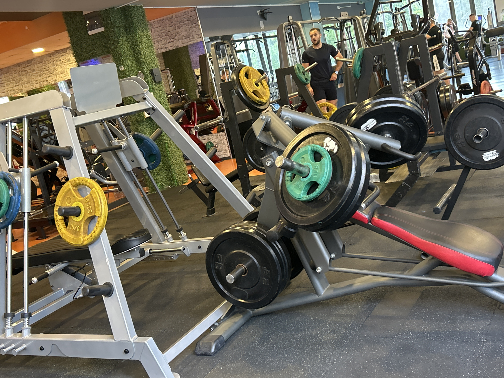 The image captures the interior of a gym in Sofia, Bulgaria, filled with various weightlifting equipment. The setting features a dark, textured floor, walls partially covered with artificial green grass and stone-like cladding, and large windows in the background letting in natural light.
In the foreground, a prominent leg press or similar machine takes up the left and center, loaded with several weight plates. Visible plates include yellow 15KG (33LB) plates and green 10KG (22LB) plates, attached to loading pins. To the right, another piece of equipment holds black 20KG (44LB) and 25KG (55LB) weight plates on storage pins. A red and black padded bench is also visible in the lower right.
In the midground, a man with a beard, wearing a black t-shirt and shorts, stands near gym machinery, looking towards the right with his mouth slightly open. He appears to be observing or pausing during a workout. Mirrors in the background reflect parts of the gym, showing other gym-goers and additional equipment, including multi-station machines and benches. Other individuals are sparsely distributed throughout the gym, engaged in various workout activities.
The scene suggests a typical daytime gym environment, with a blend of natural light from the windows and artificial overhead lighting illuminating the space. The gym appears well-maintained and equipped for strength training.
