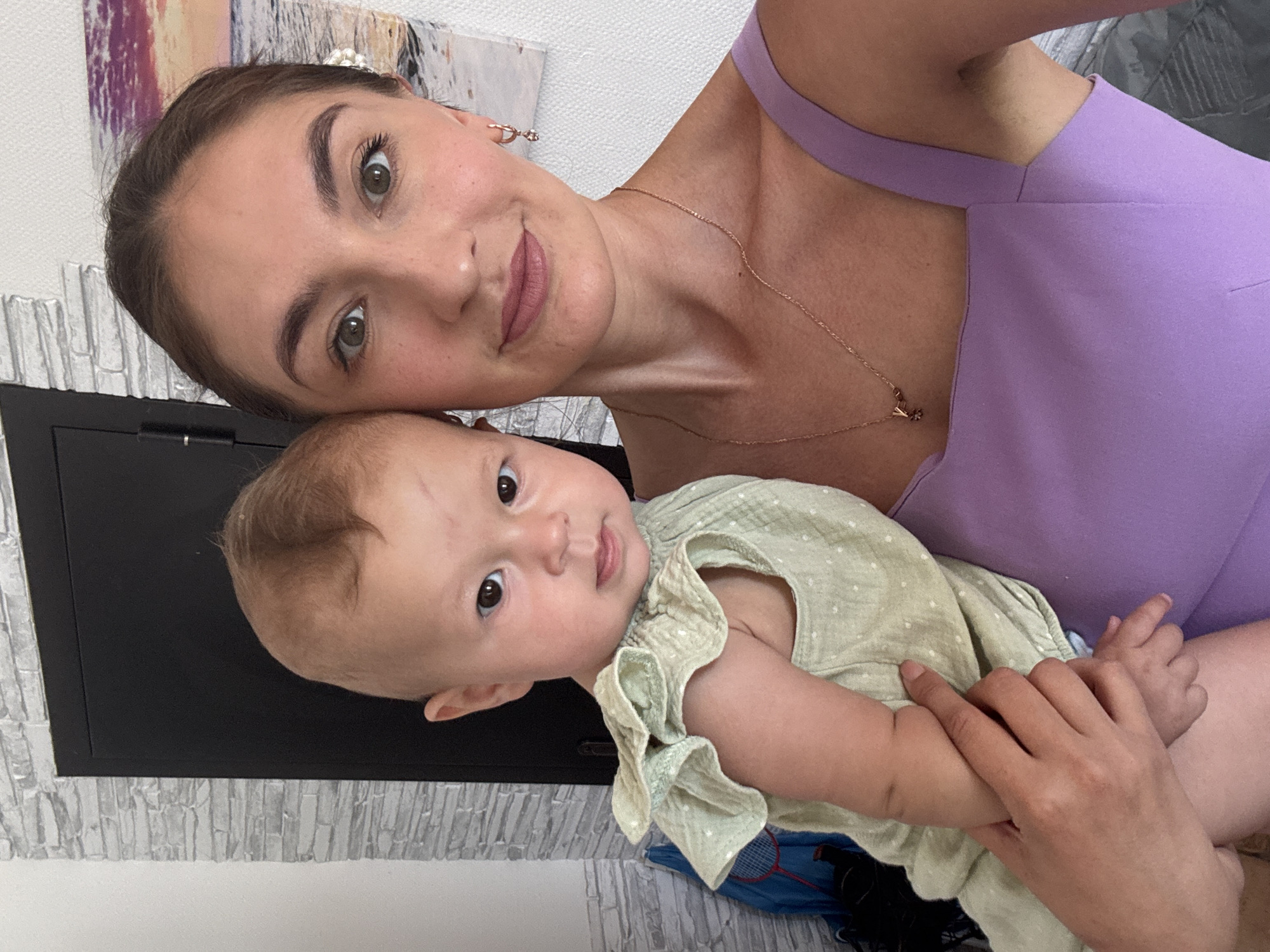 This media file shows a woman holding a baby. The woman is wearing a purple dress and a delicate gold necklace. The baby is wearing a light green outfit with small white polka dots. They appear to be indoors, with a patterned wallpaper in the background. The overall mood is one of affection and closeness between the woman and the baby. The lighting suggests it is daytime.