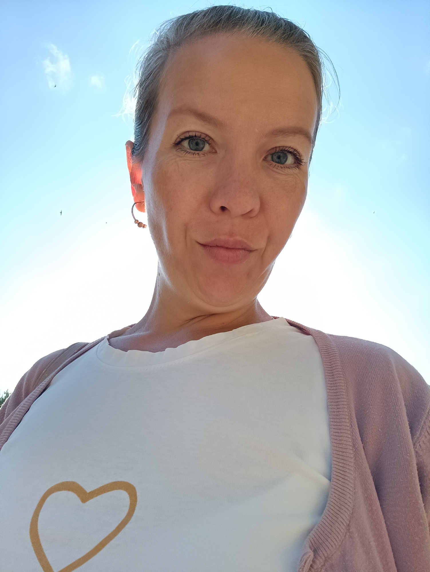 An outdoor, low-angle, close-up, selfie-style photograph shows a fair-skinned woman with light brown hair pulled back from her face. She is looking at the camera with a slight smile and her head is tilted upwards, obscuring the top of her head. The bright blue sky with a few wispy white clouds and two small birds in flight is visible behind her. She is wearing a white t-shirt with a large, outlined gold heart printed on the front and a light pink cardigan. A silver hoop earring with small beads is visible in her left ear. The background is out of focus. The image was taken on July 14, 2023, at an outdoor location in Chaikovsky, Russia.