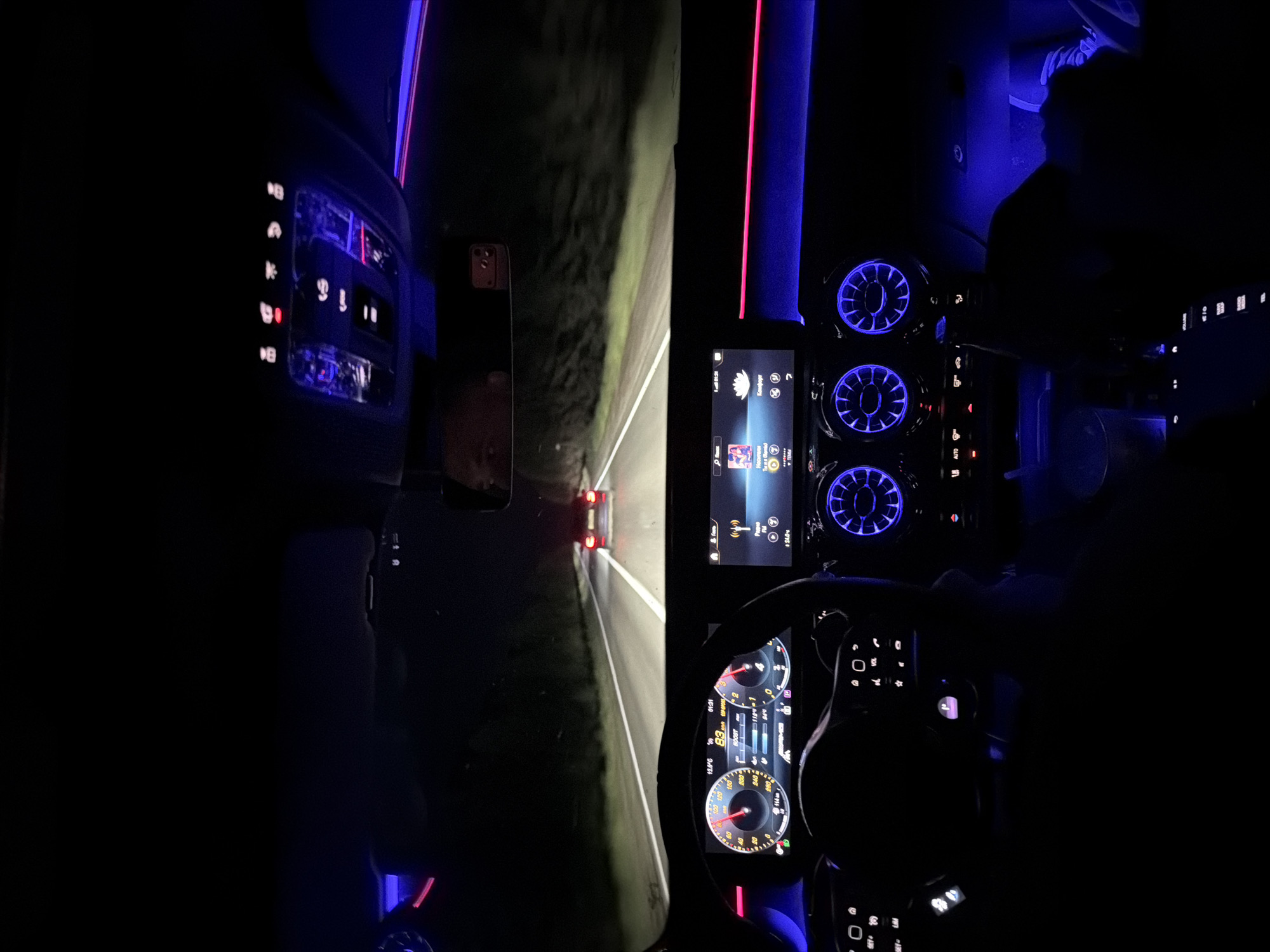 This image shows the interior of a car at night. The dashboard, steering wheel, and central console are visible, illuminated by blue and red lights. A display screen is lit up, showing various car functions. The driver's hands are on the steering wheel, and a road is visible through the front windshield. The location is indicated by a sign "комфорт" and a radio station listed at 94.0.
