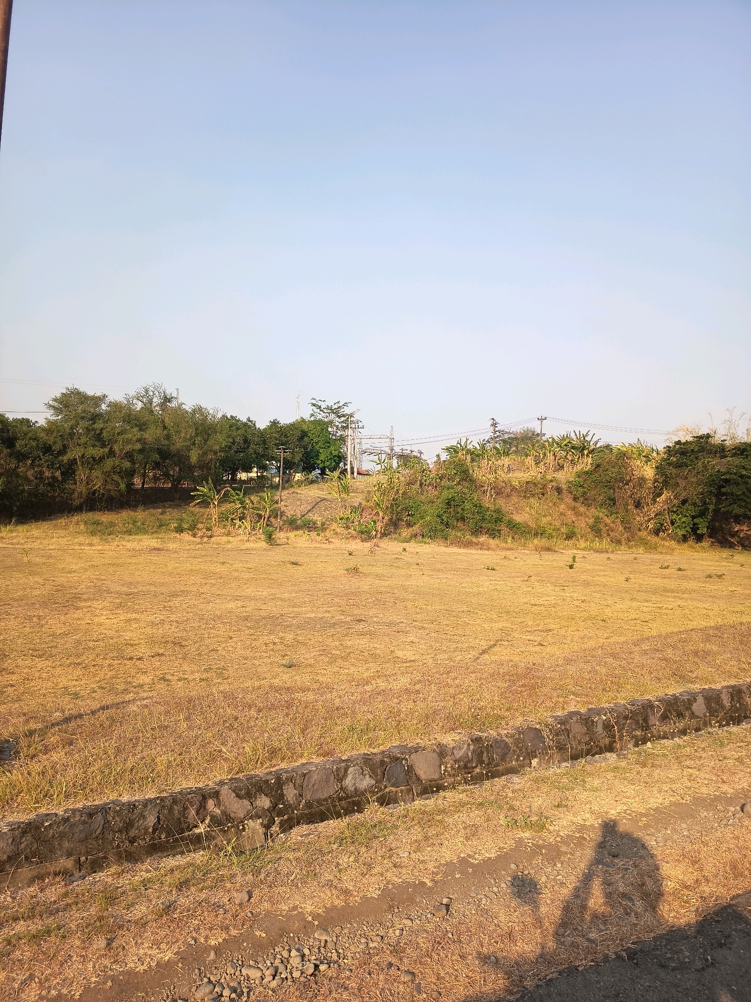The image depicts a dry, grassy field in Kalangbancar, Indonesia. The field is mostly barren with patches of dry, yellow grass. A low stone wall runs along the edge of the field, separating it from a gravelly path. In the background, there is a line of trees and vegetation, and further back, electrical transmission towers and power lines are visible. The sky is clear and blue. Two human shadows are cast on the ground in the foreground, indicating the presence of at least two people out of frame.