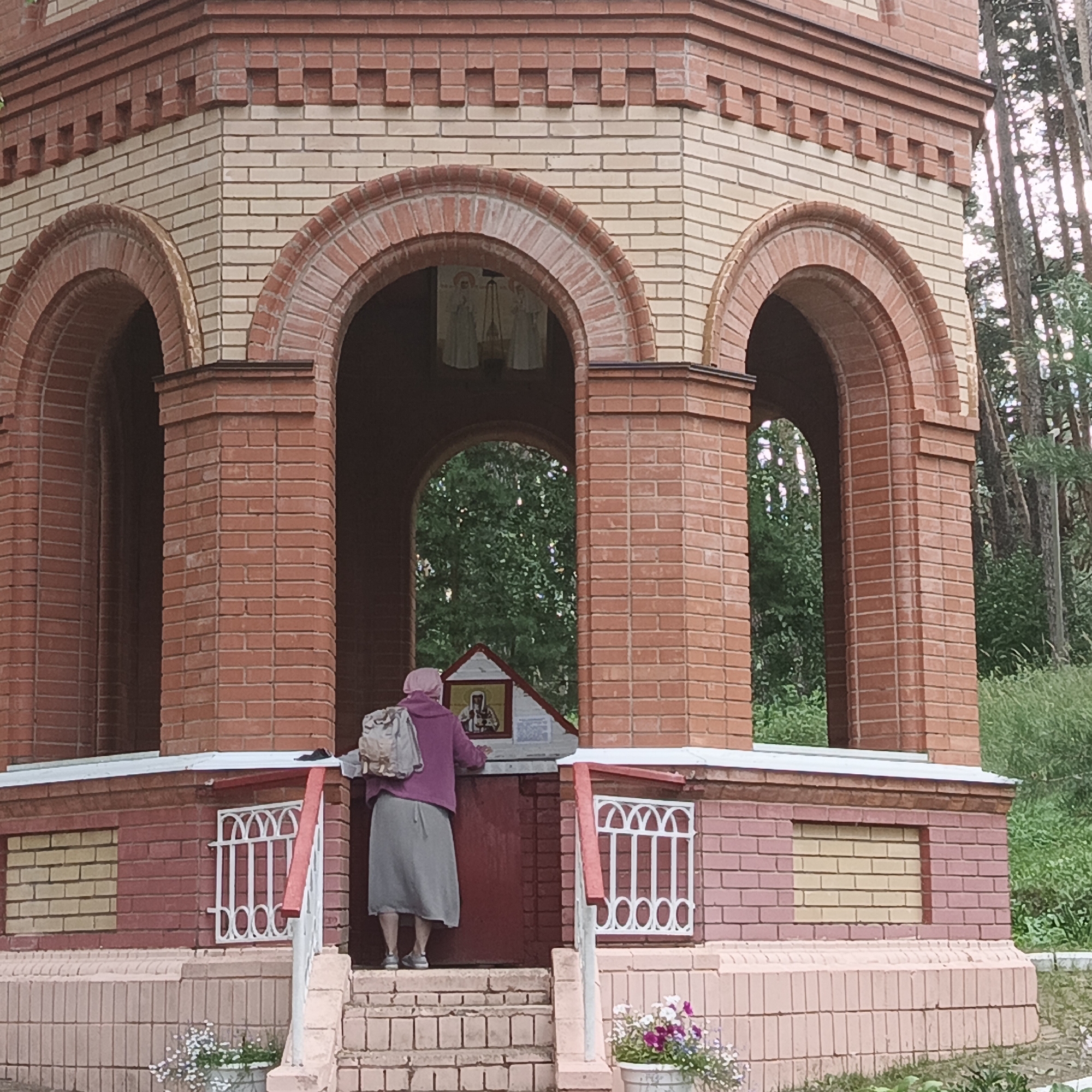 In Nikolo-Beryozovka, Russia, a person wearing a pink headscarf and a purple hooded jacket over a grey skirt stands on steps in front of a small, red-brick structure with arched openings. They have a backpack on and appear to be looking at an icon displayed on a small roofed structure attached to the building. The building's architecture features alternating bands of red and beige bricks, with decorative brickwork along the top edges of the arches. White metal grates are visible on the lower part of the structure, and potted flowers are placed near the base. The background consists of lush green trees, suggesting a natural, outdoor setting.