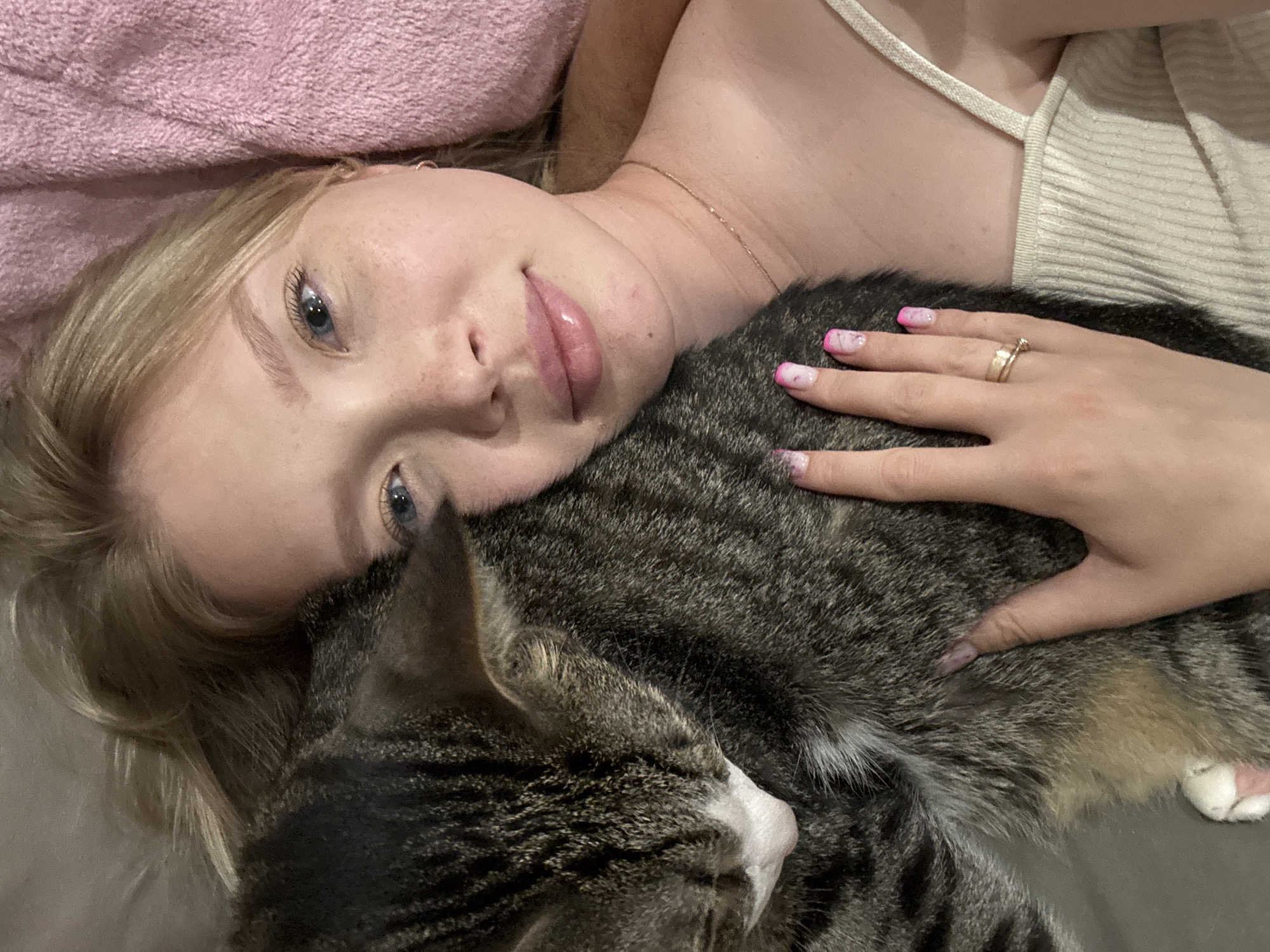 In Chaikovsky, Russia, a young woman with blonde hair and freckles is captured in a selfie, embracing a tabby cat. The woman is wearing a light-colored tank top and a thin gold necklace. She has bright blue eyes and glossy pink lips. Her fingernails are painted with a pink and white marble design. A ring is visible on her right ring finger. The cat is nestled against her face, its fur a mix of gray, black, and white. The background appears to be a soft surface, possibly a blanket or bed, which is pink in color on the right side of the frame. The lighting is dim, suggesting an indoor setting, likely a bedroom.