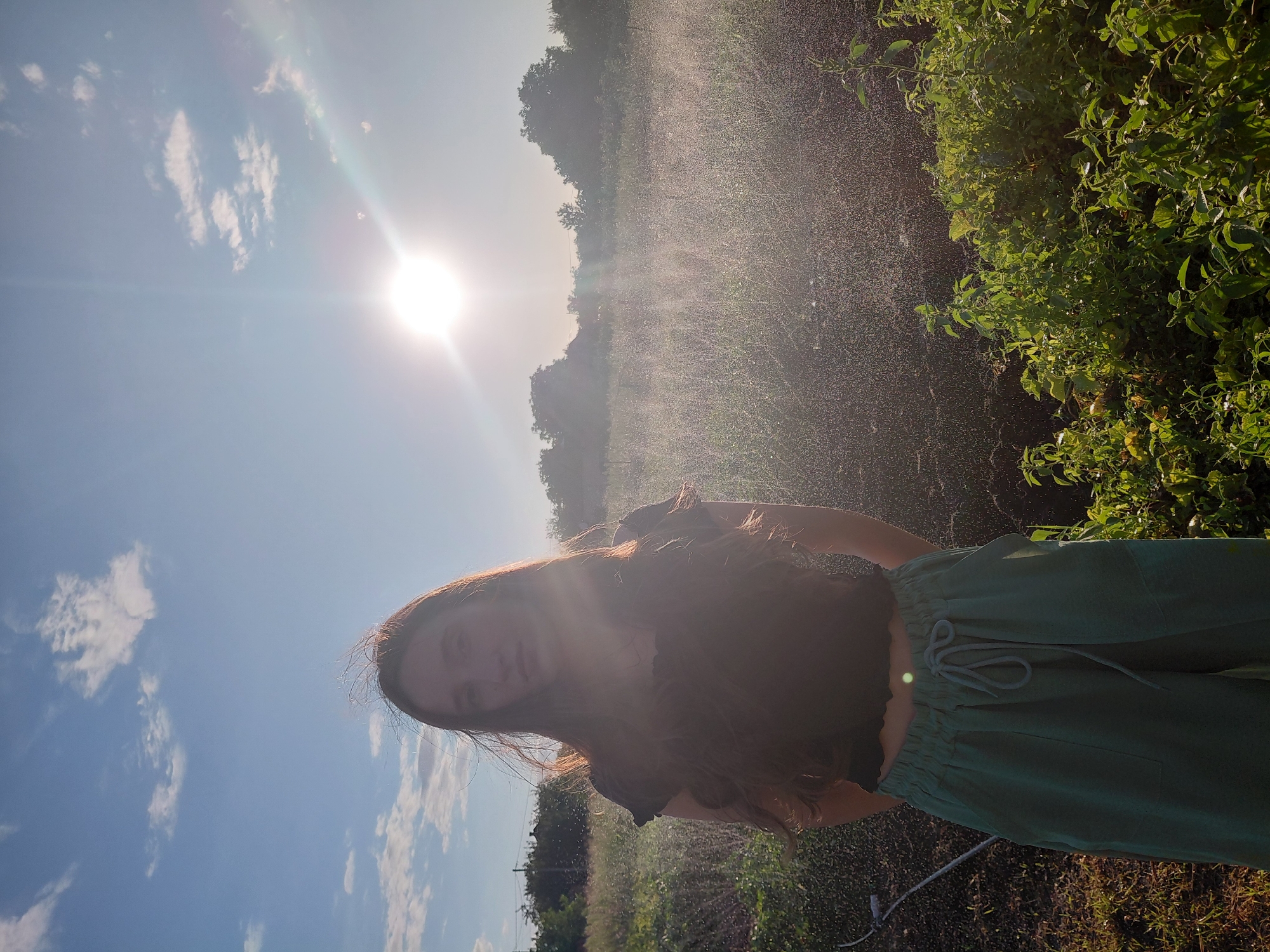 A young woman with long, brown hair stands outdoors in a garden at dusk. She is wearing a dark, cropped top and loose, green pants. The sun is setting behind her, creating a lens flare and casting a warm glow on the scene. Water is being sprayed in the background, creating a misty effect over the plants and soil. The garden appears to be in a rural or suburban setting, with trees and a building visible in the distance.