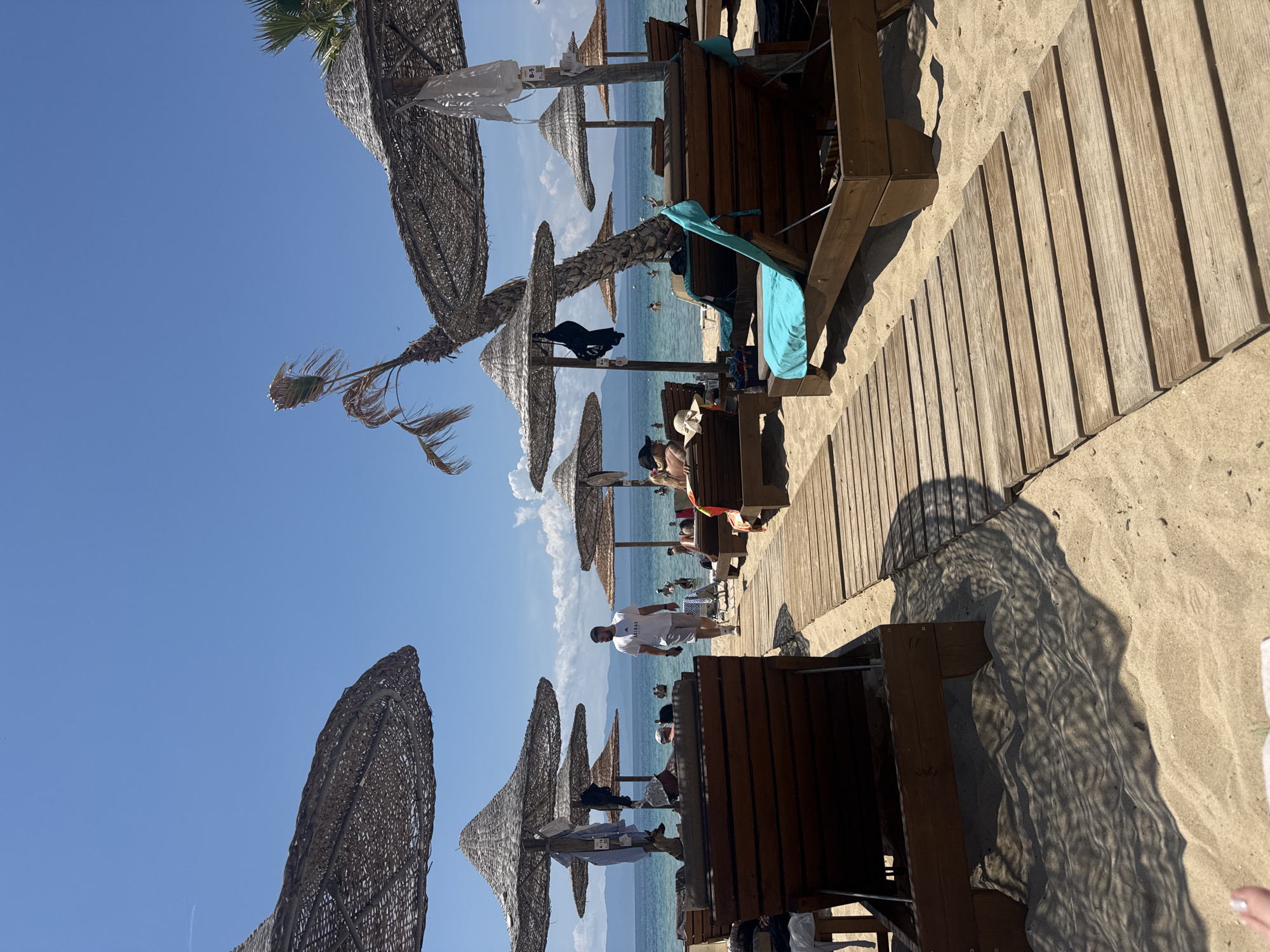 This is a daytime, eye-level shot of a beach at Ofrynio Beach, Greece. The sand is light brown, and a wooden boardwalk runs diagonally from the lower left to the upper right. Several wooden lounge chairs, some with light blue towels, are positioned along the boardwalk and on the sand. Woven straw umbrellas provide shade over some of the lounge chairs. In the background, people are swimming and relaxing in the clear blue ocean. Distant mountains can be seen on the horizon under a bright blue sky. A few palm trees are visible, one of which is leaning precariously. Several items of clothing are hanging on the umbrellas. The scene depicts a typical beach resort atmosphere.