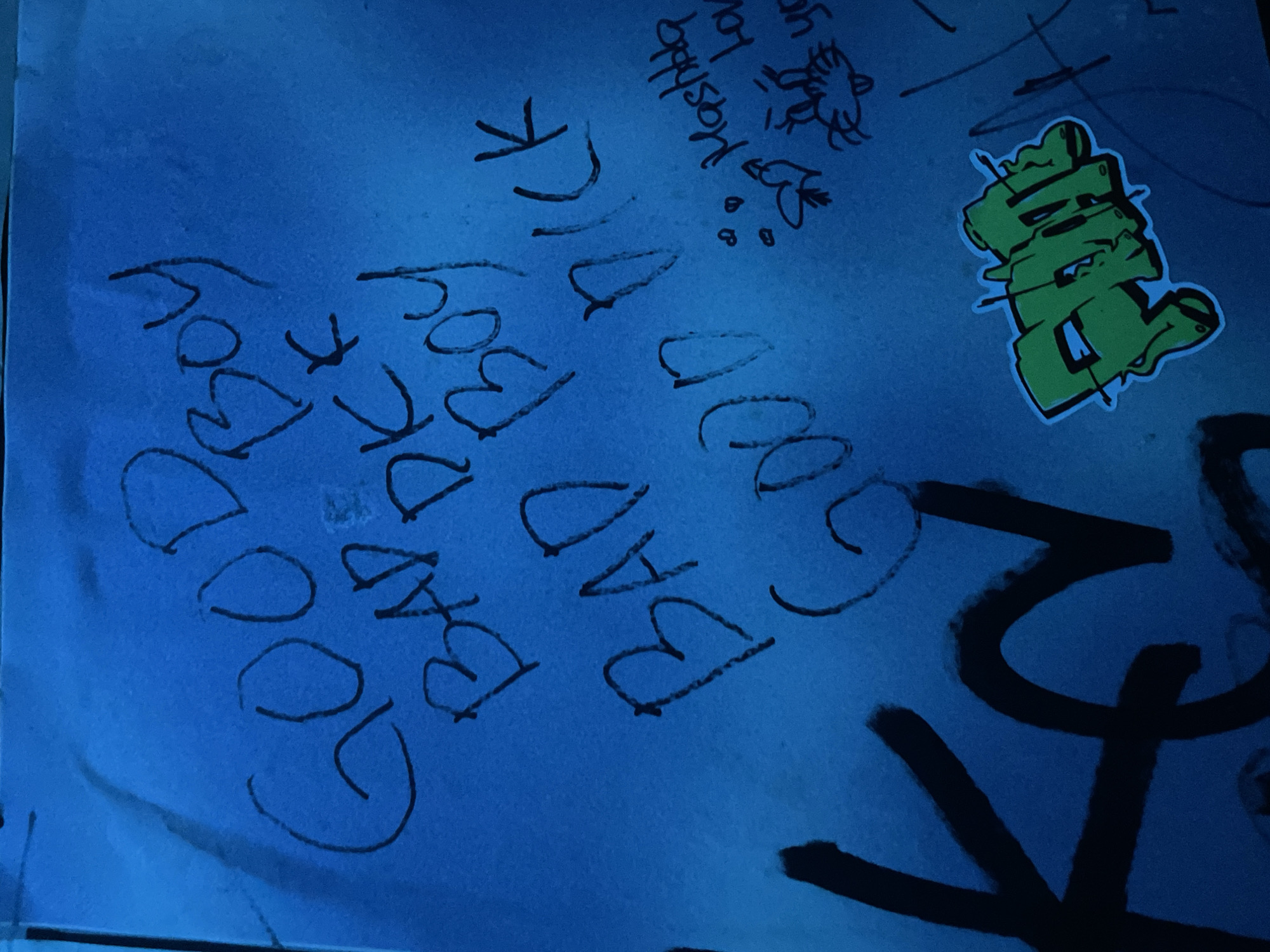 The image shows a close-up of graffiti on a light blue surface. The graffiti includes the phrases "GOOD BOY" and "BAD BOY LUCK" written in simple, dark ink lettering. There is also a green sticker with an abstract design. Above "BAD BOY LUCK" is the phrase "Yoshka lov". Small drawings of animals and hearts accompany the writing. Large black spray-painted lines are visible on the bottom right of the surface. There is no information about the location in the city of North Kuta, Indonesia in the image.