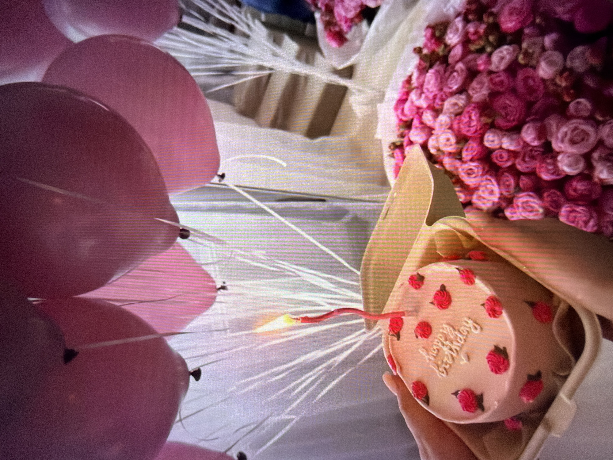 In Beșalma, Moldova, a pink cake with "happy birthday" written in white icing and decorated with pink roses sits in a tan holder. A lit red candle stands on the cake. To the right of the cake, a bouquet of pink roses is visible. Pink balloons float in the background. A hand is holding the cake holder.