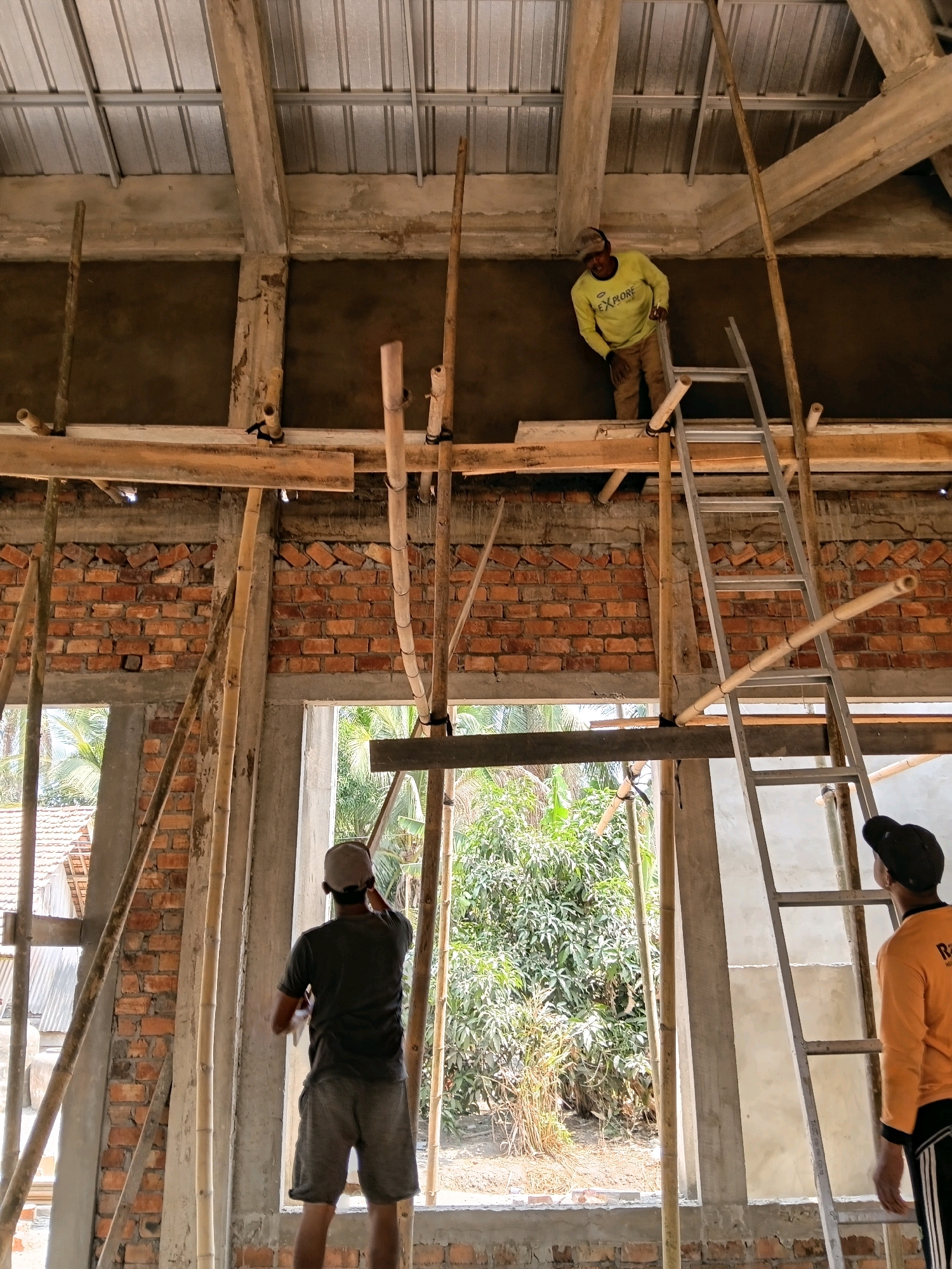 In Margo Rukun, Indonesia, a construction site is depicted under a corrugated metal roof supported by concrete beams and bamboo scaffolding. Three men are present. In the foreground, a man wearing a grey t-shirt and shorts, and a baseball cap faces away from the camera, working on a wall. To the right, another man in an orange long-sleeved shirt and black and white striped pants looks up, standing near a metal ladder. Higher up on the scaffolding, a man in a yellow long-sleeved shirt and brown pants is also visible, appearing to work on the wall. The walls are constructed of red brick, and windows offer views of tropical foliage and houses.