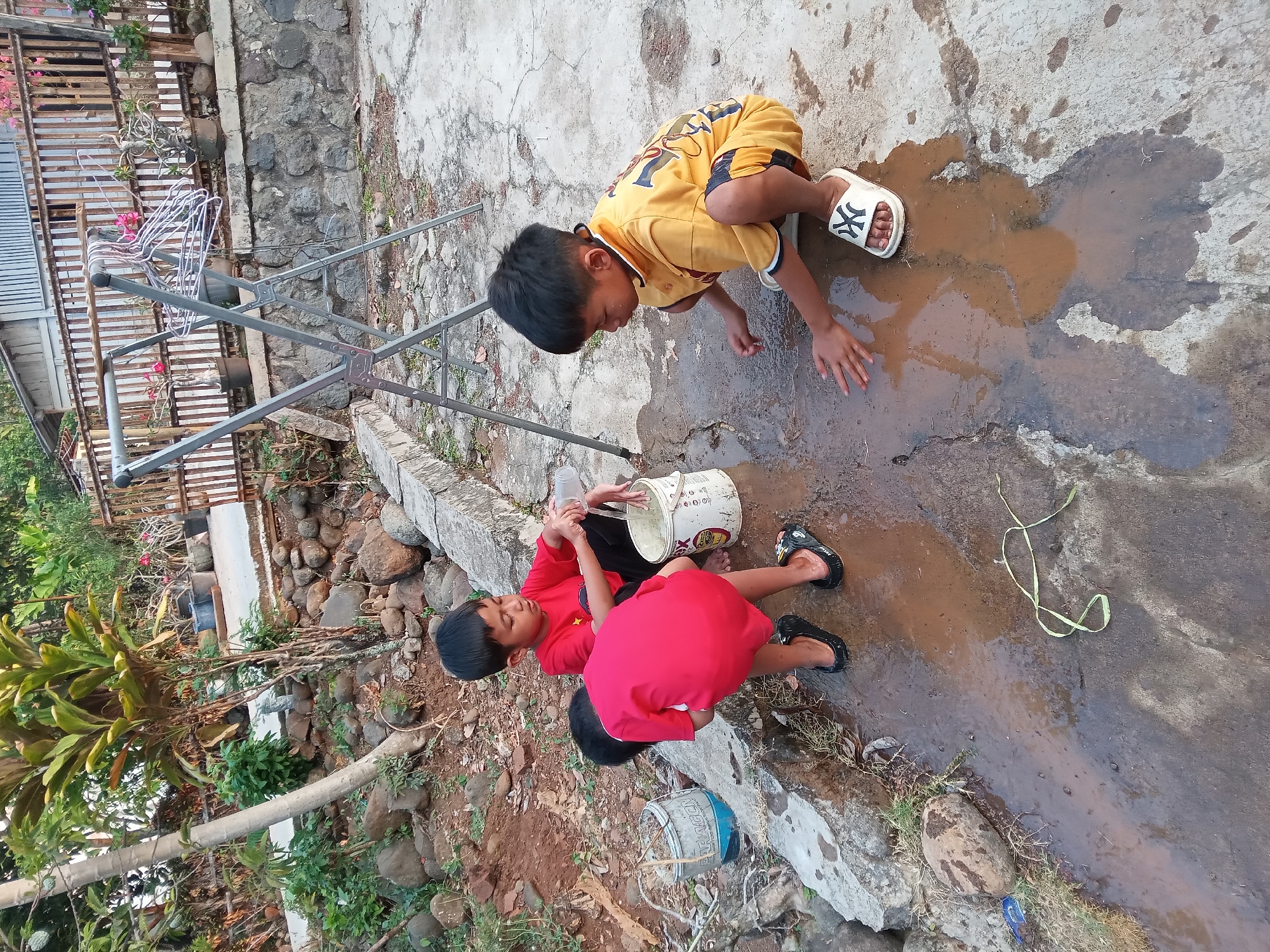 The image depicts a rural scene in Kedunggudel, Indonesia, where three young boys are playing in a muddy puddle on a concrete surface. In the foreground, two boys are interacting with the water. One boy, wearing a red t-shirt and black shorts, is bending over near a white bucket and appears to be pouring water from a clear plastic bottle into the puddle. Another boy, to his right, is also in a red t-shirt and black shorts, with his back partially to the camera, and is playing in the water. A third boy, on the right side of the frame, is wearing a yellow t-shirt and shorts and is squatting down, actively playing with the mud and water. He is wearing white sandals with a black "NY" logo.
The environment suggests a backyard or outdoor area with a rough concrete floor. Rocks and dirt are visible on the left side, along with some overgrown vegetation. In the background, a wooden fence encloses an area with some potted plants and a metal drying rack with clothes hangers. A simple building with a corrugated roof is visible behind the fence. The lighting suggests it is daytime, possibly late afternoon, with soft shadows indicating the sun's position. The overall impression is one of children engaged in simple outdoor play in a modest, rural setting.